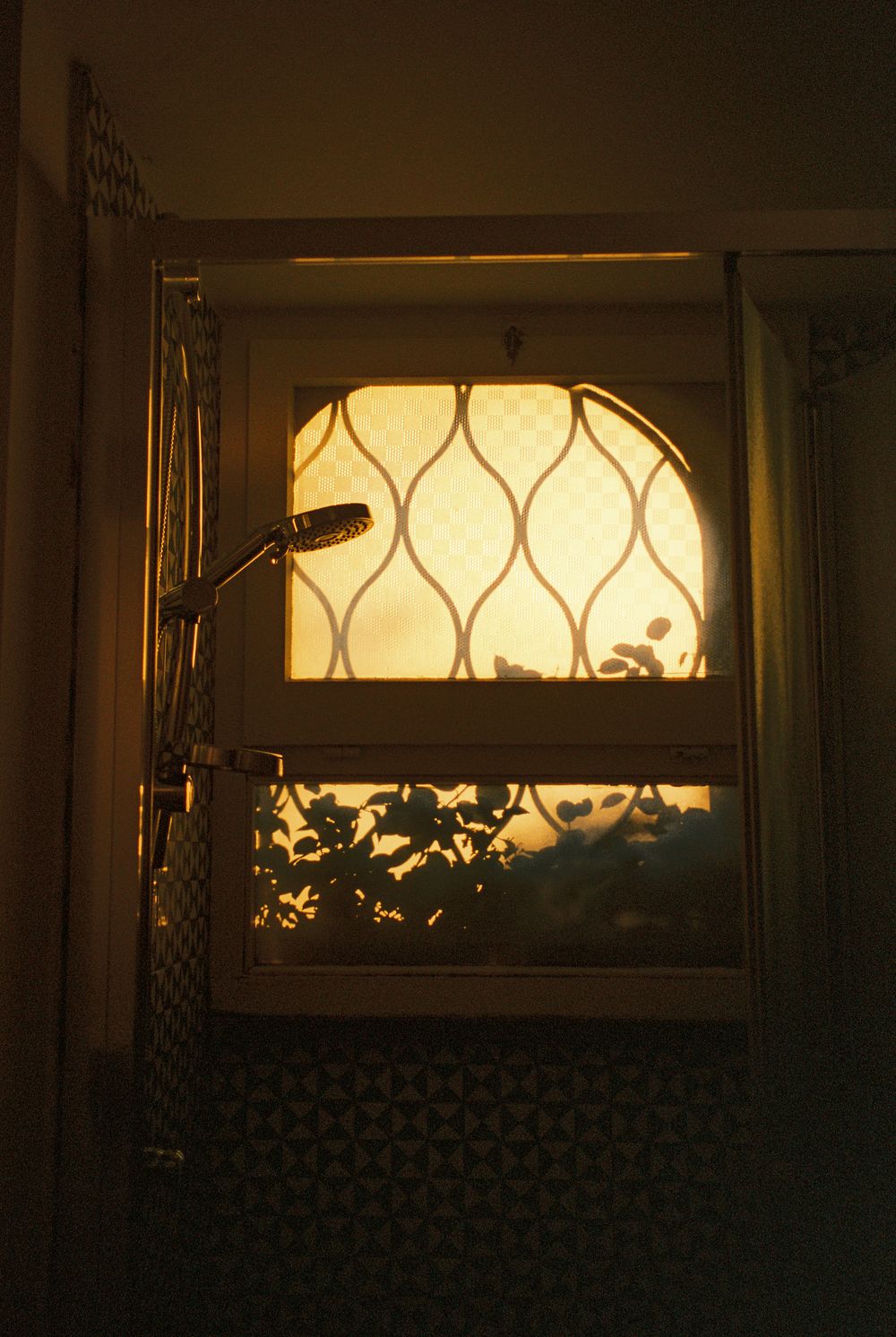 Golden sunset light glows through a decorative lattice bathroom window with silhouetted plants and a shower head.