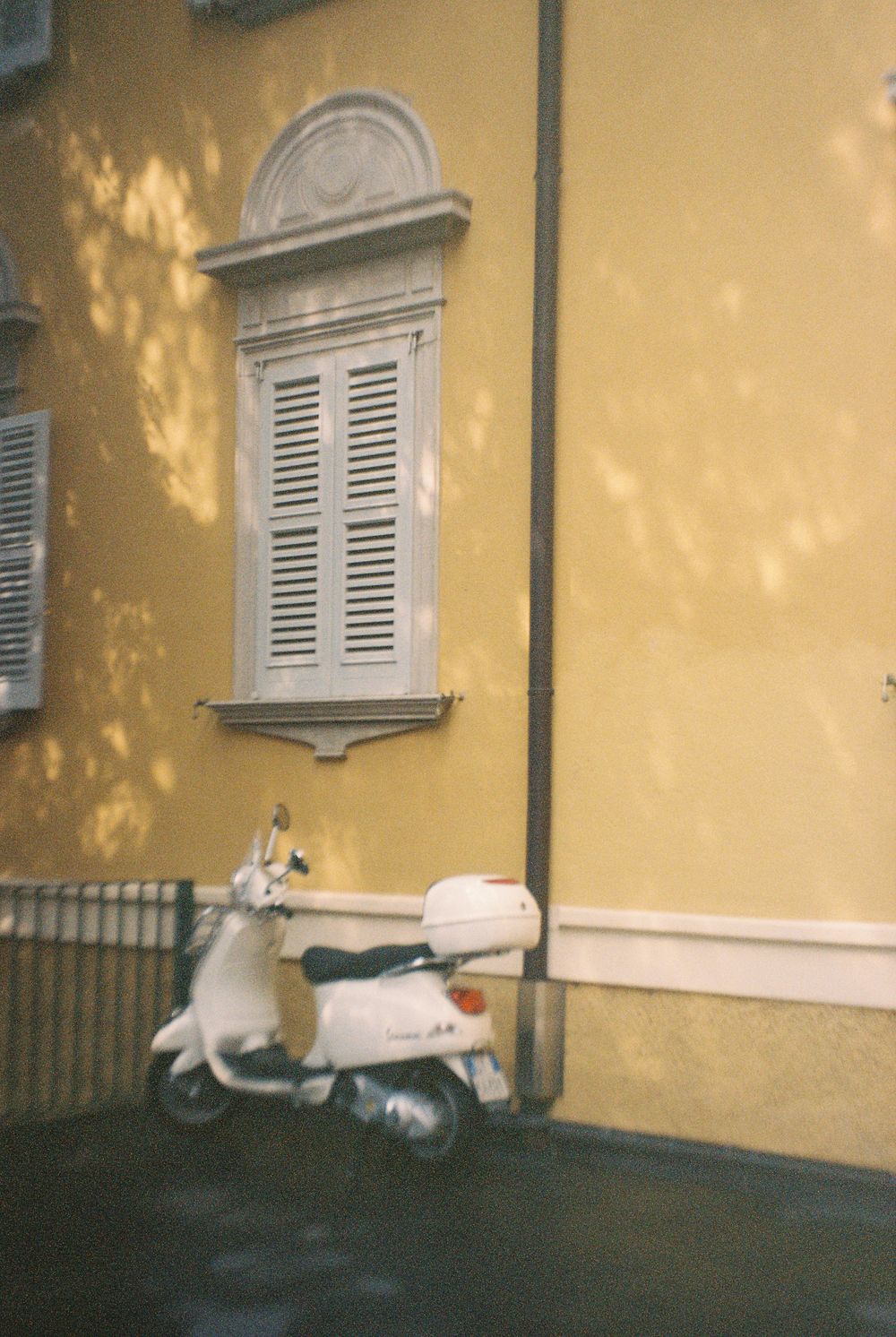 White Vespa scooter parked against a yellow building with shuttered window in Europe.