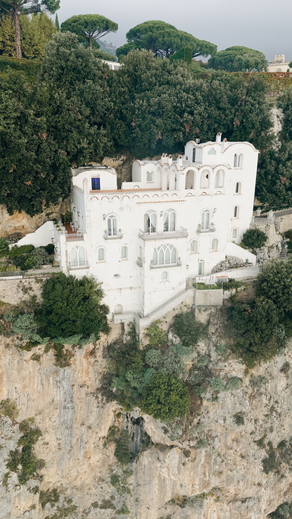 Aerial view of grand white villa perched on rocky cliffside surrounded by lush Mediterranean trees.