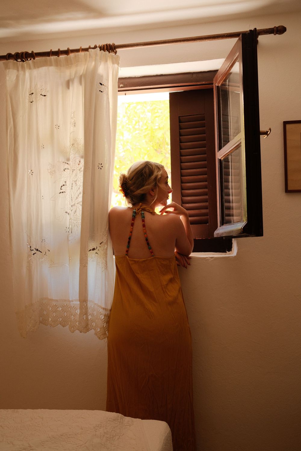 Woman in mustard dress gazing out open wooden shuttered window with lace curtains in warm sunlit room.