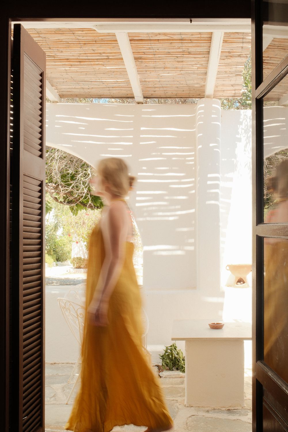 Blurred woman in golden maxi dress walking through open doors of a sunlit Mediterranean villa.