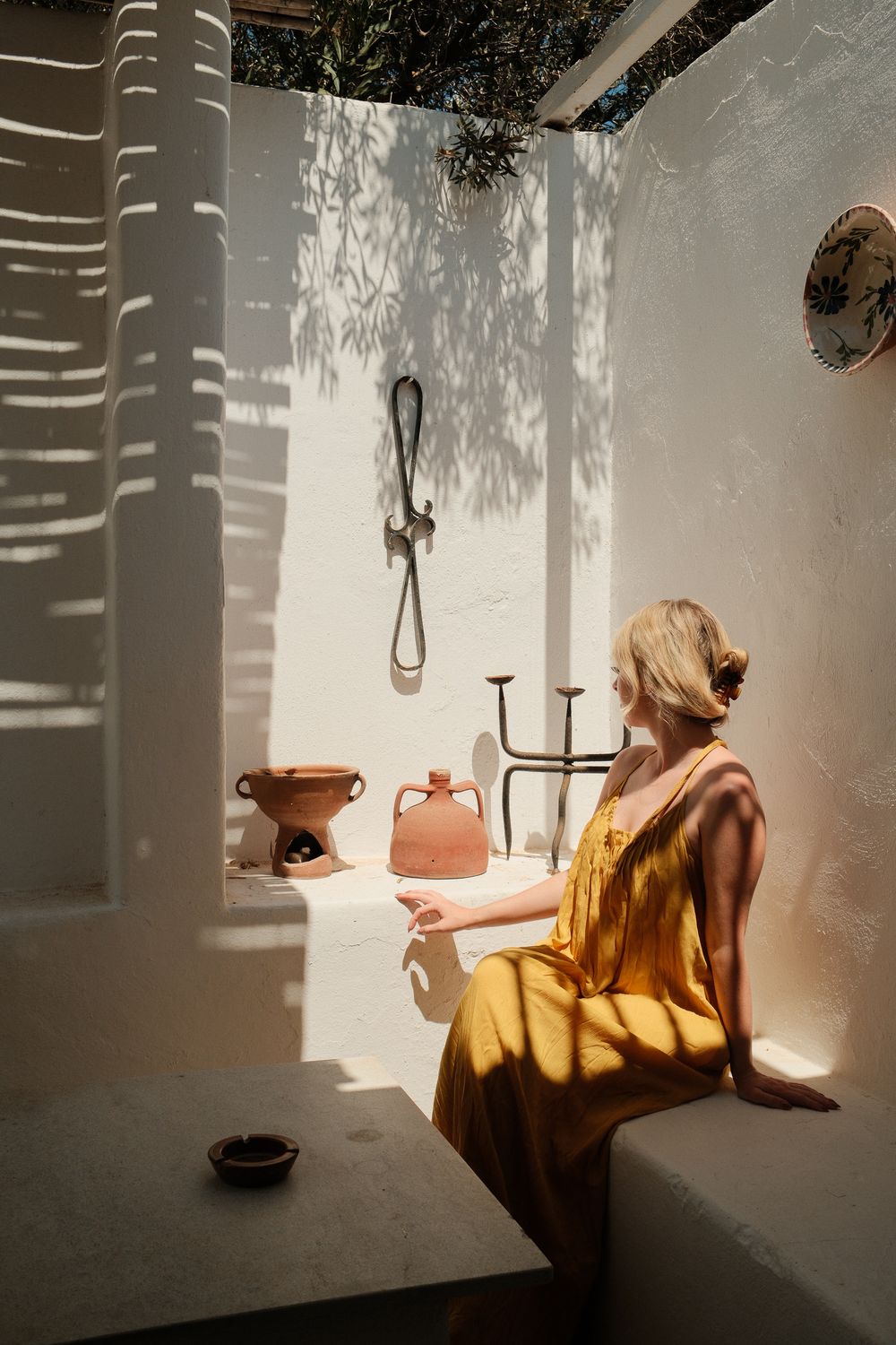 Woman in yellow dress sitting by white Mediterranean wall with terracotta pottery and dappled sunlight shadows.