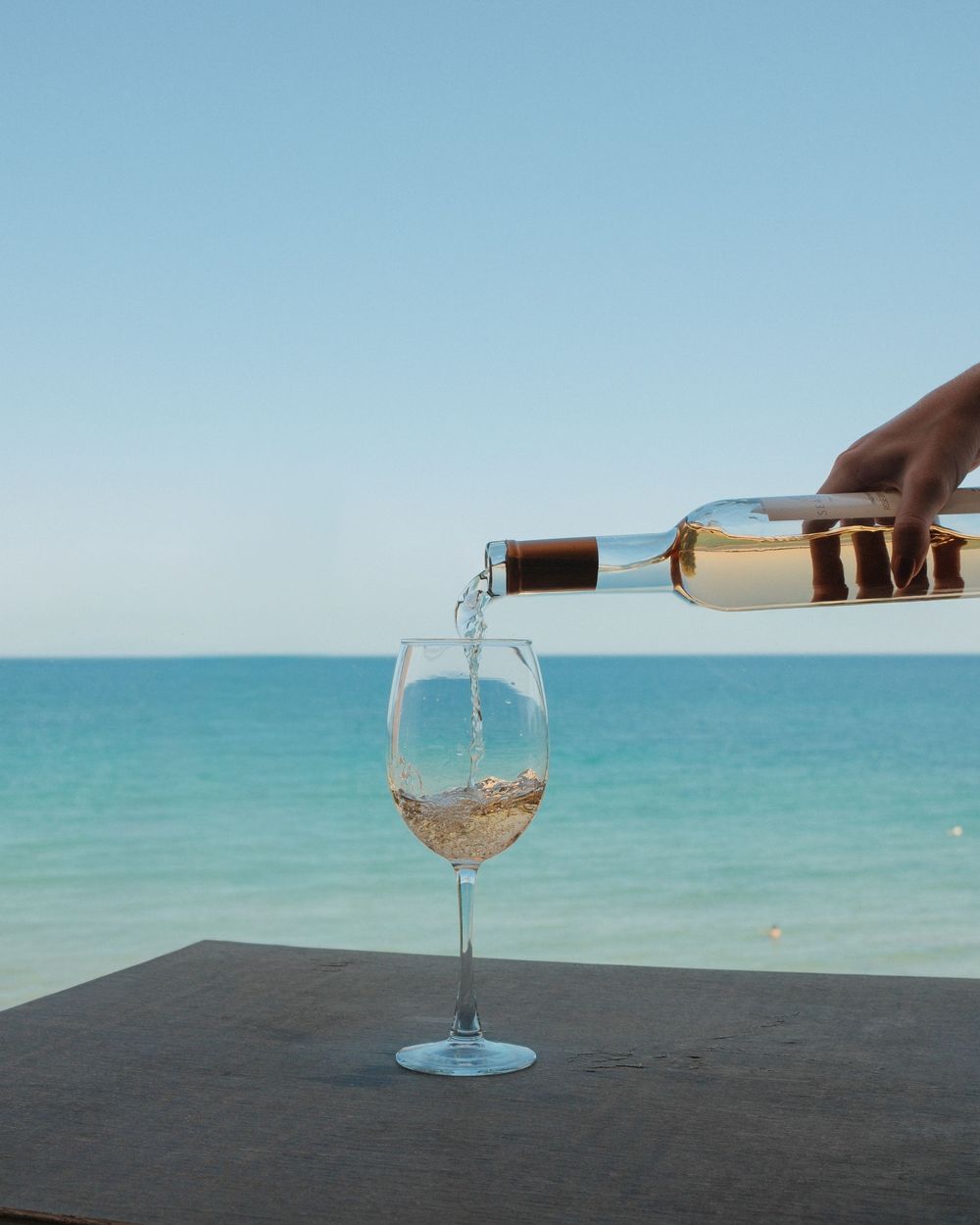 Hand pouring white wine into a glass on a table with a stunning turquoise ocean and clear blue sky in the background.