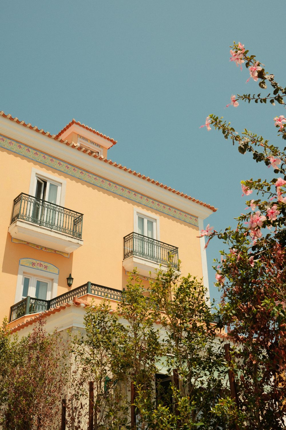 Yellow Mediterranean building with wrought iron balconies and pink flowering tree against clear blue sky.