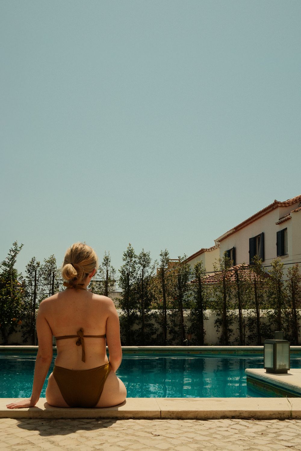 Blonde woman in a brown bikini sitting by a sunny outdoor pool with trees and a white villa in the background.