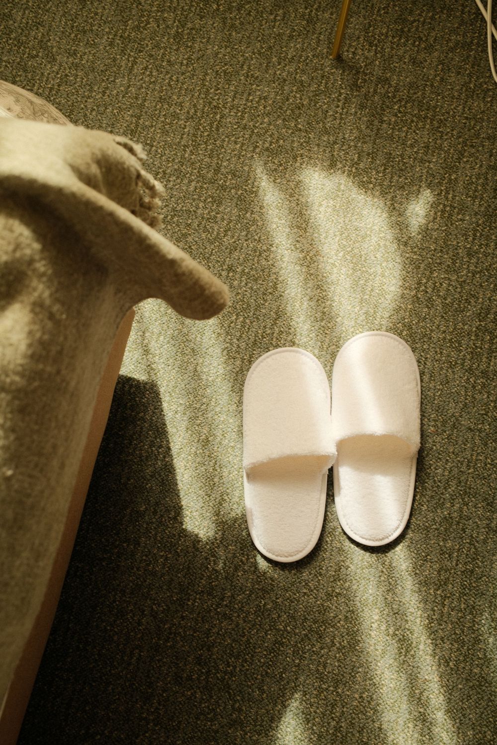 White hotel slippers placed on green carpet beside a bed in warm sunlight.