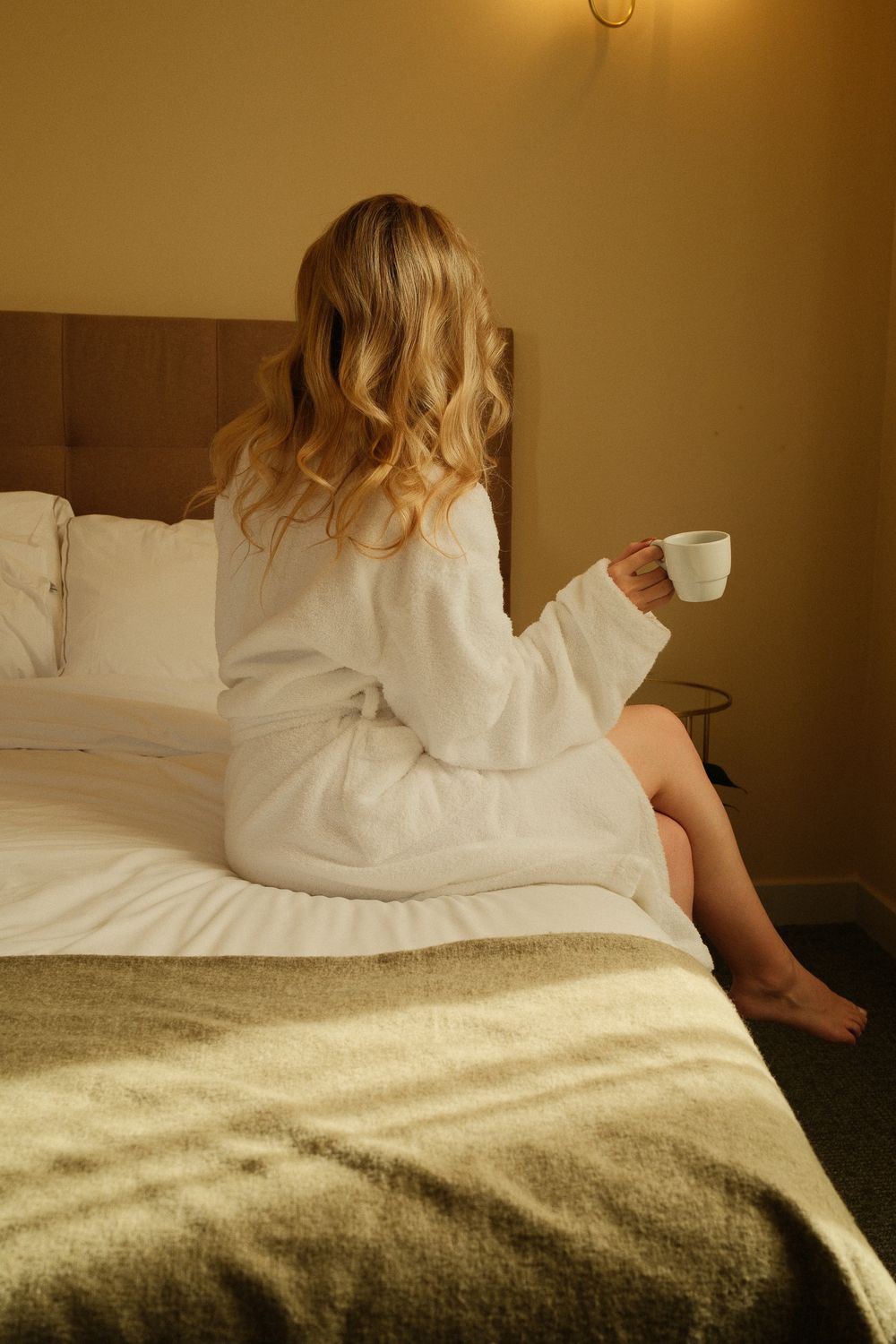 Blonde woman in white bathrobe sitting on hotel bed holding a white coffee cup in warm morning light.