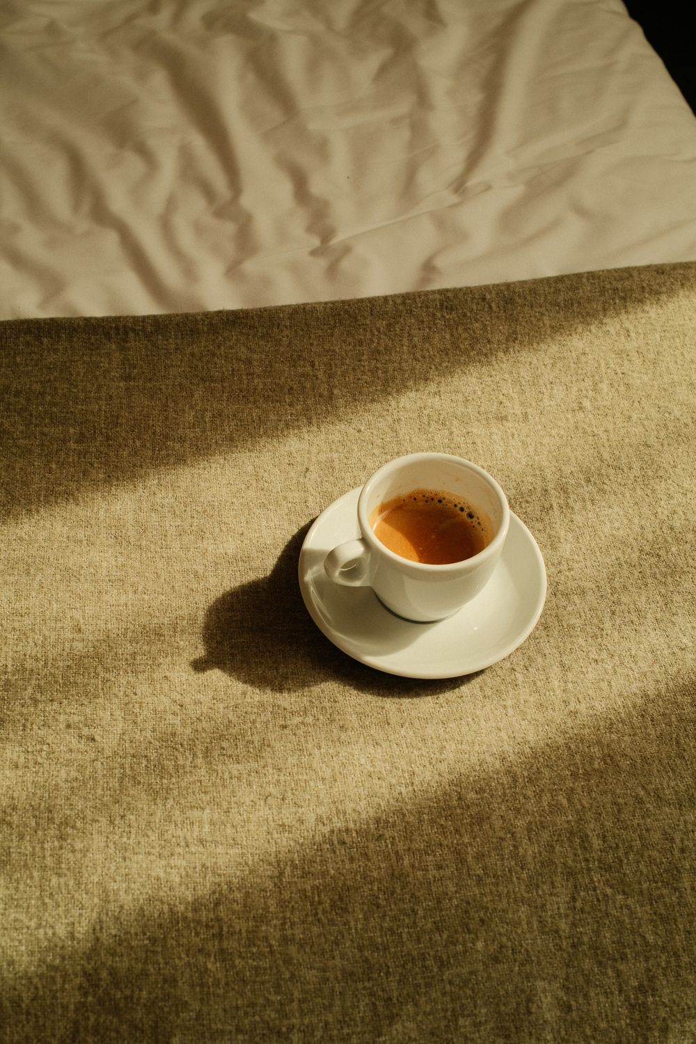 A white espresso cup on a saucer sits on a textured beige surface, bathed in warm sunlight casting soft shadows.