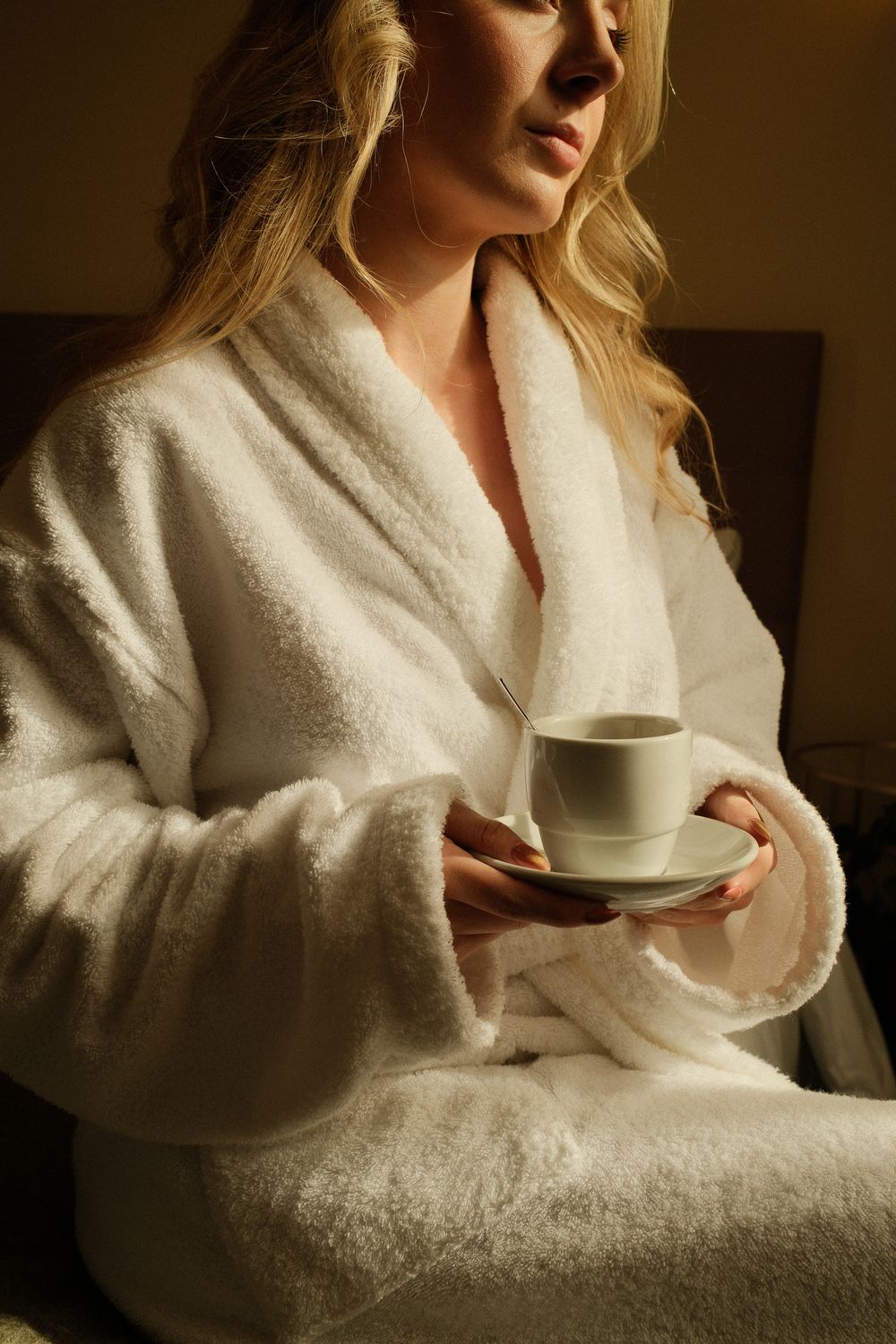 Blonde woman in white fluffy bathrobe holding a cup and saucer, enjoying a morning coffee in a cozy room.