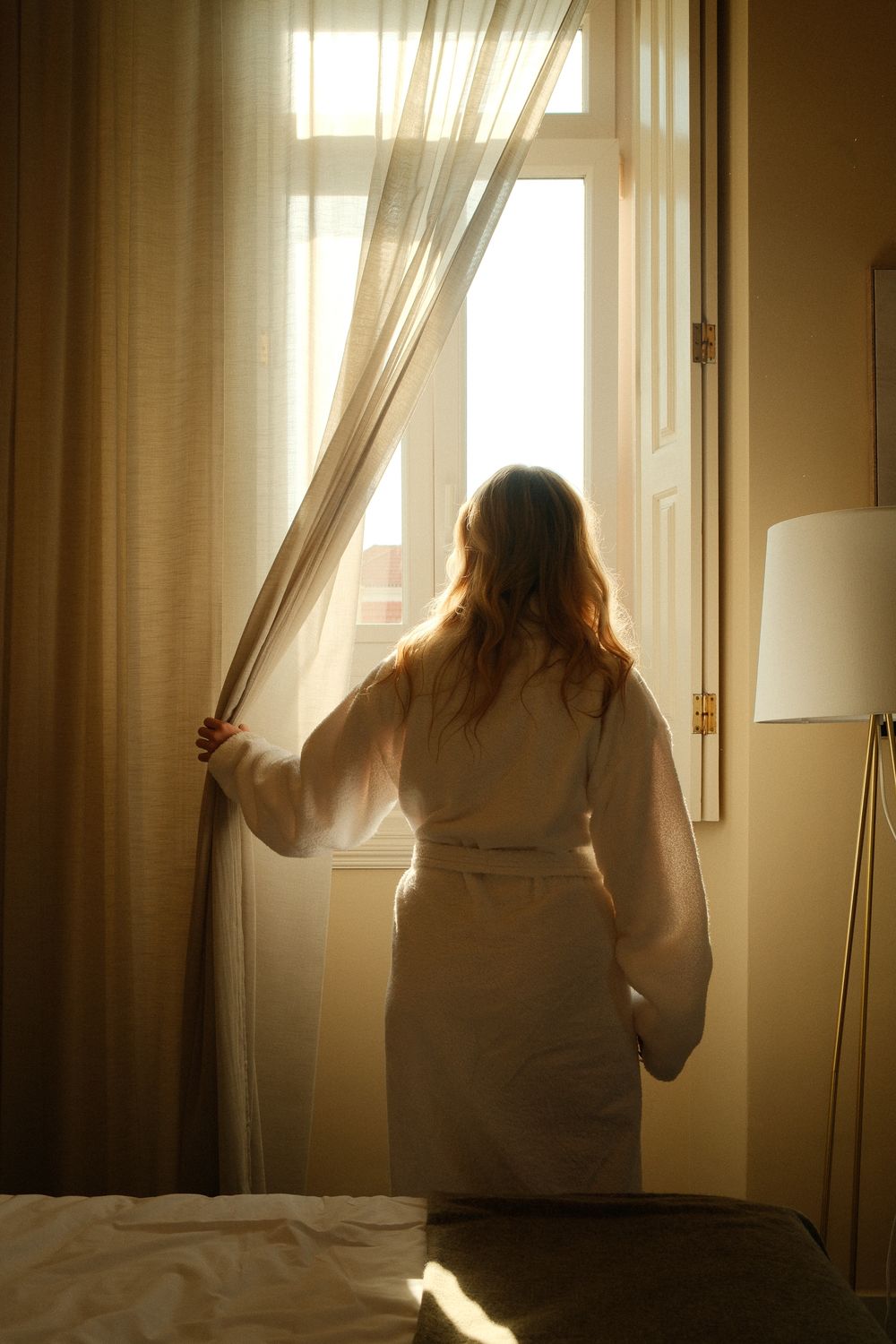 Woman in white bathrobe opening sheer curtains to let morning sunlight into a warmly lit hotel bedroom.