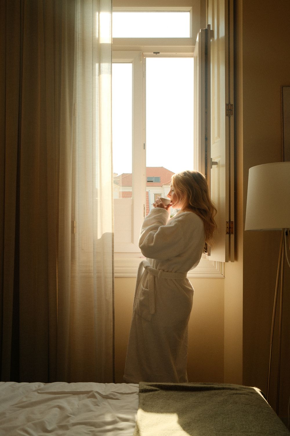 Woman in white bathrobe standing by open window in golden morning sunlight in a cozy bedroom.
