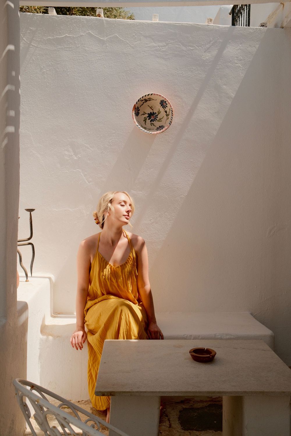 Woman in yellow dress sitting on white Mediterranean wall in sunlight.