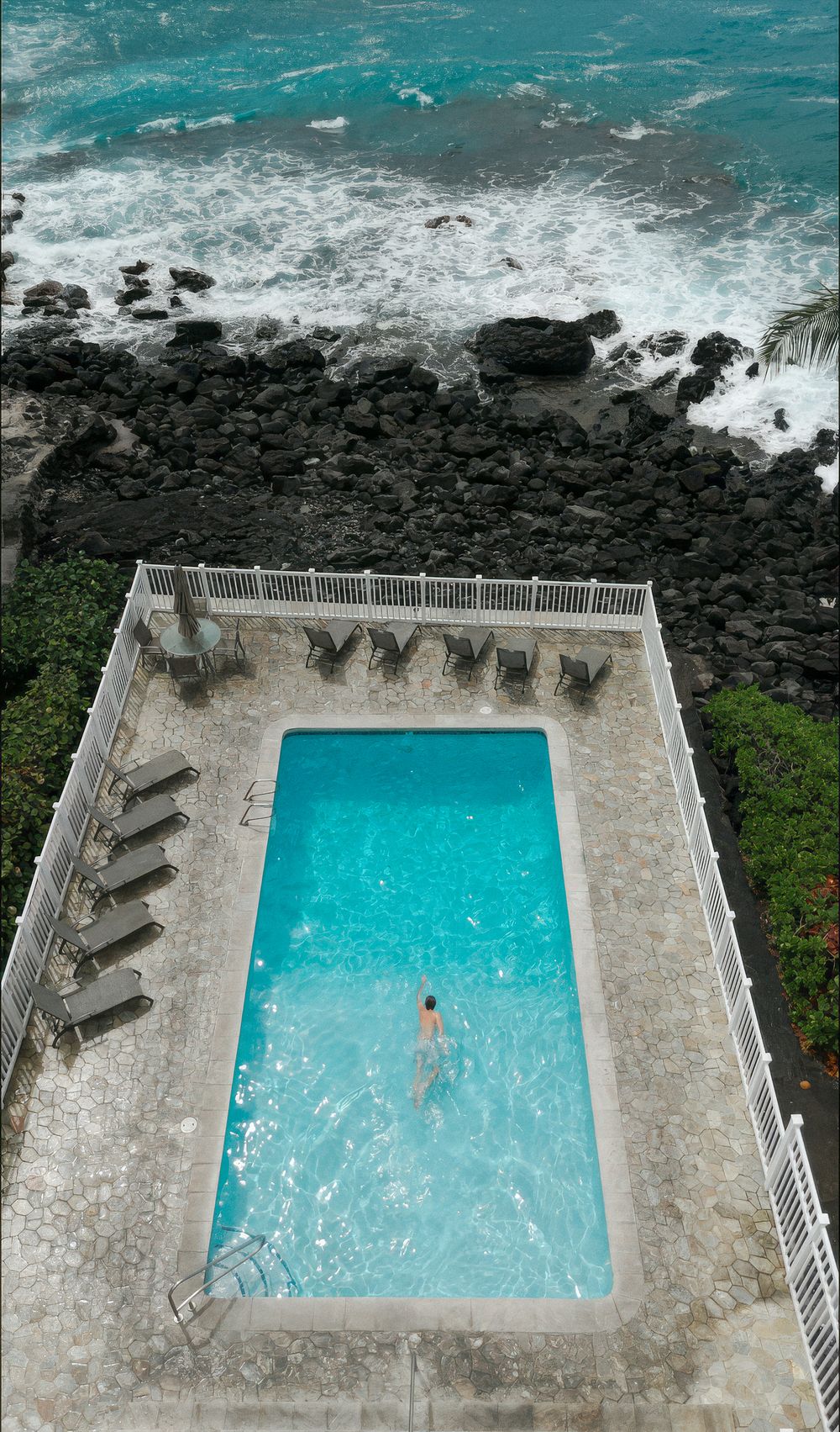 Aerial view of a rectangular pool beside rocky volcanic coastline with turquoise ocean waves.