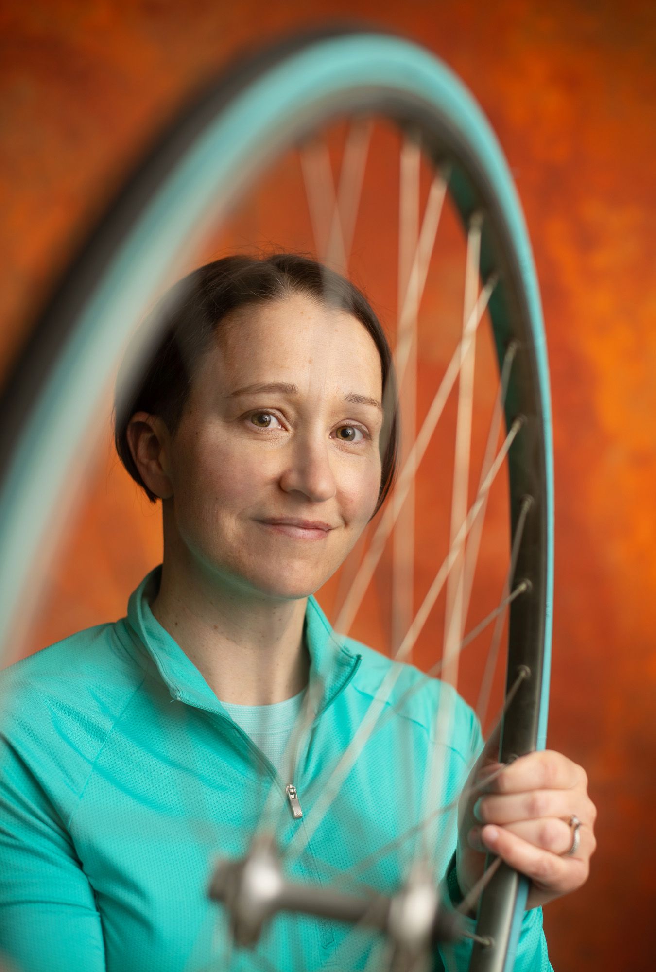Woman in teal jacket holding a bicycle wheel with spokes visible against a warm orange background.