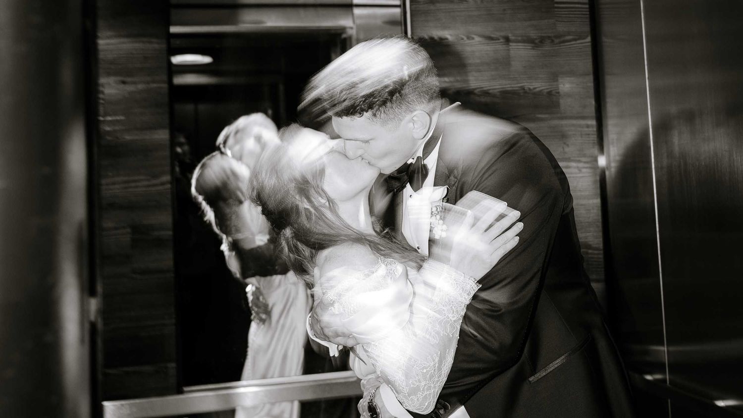Bride and groom share romantic kiss under grand staircase with marble columns in elegant wedding venue.