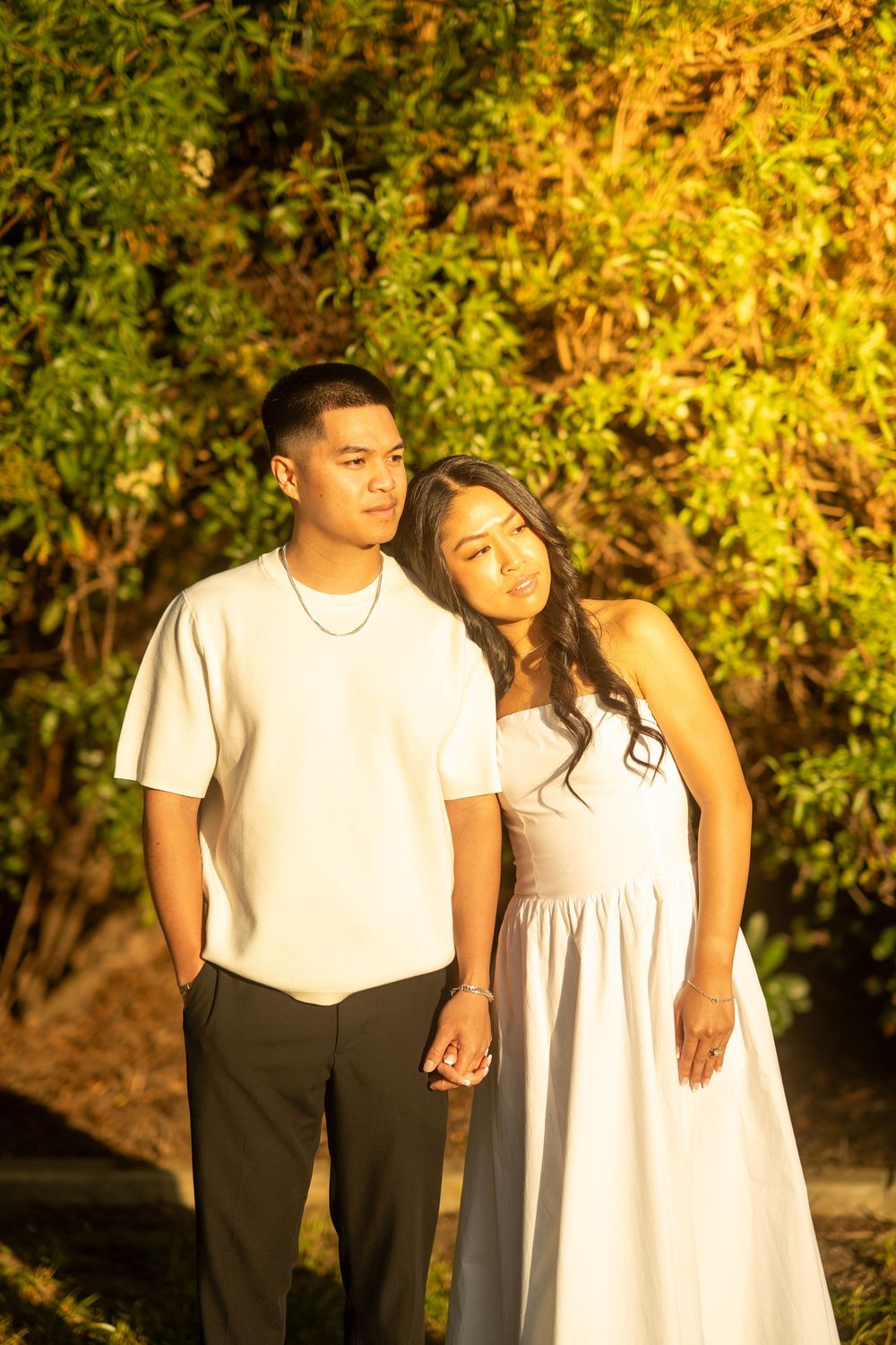 Young couple in white outfits holding hands at golden hour with lush green trees in background.