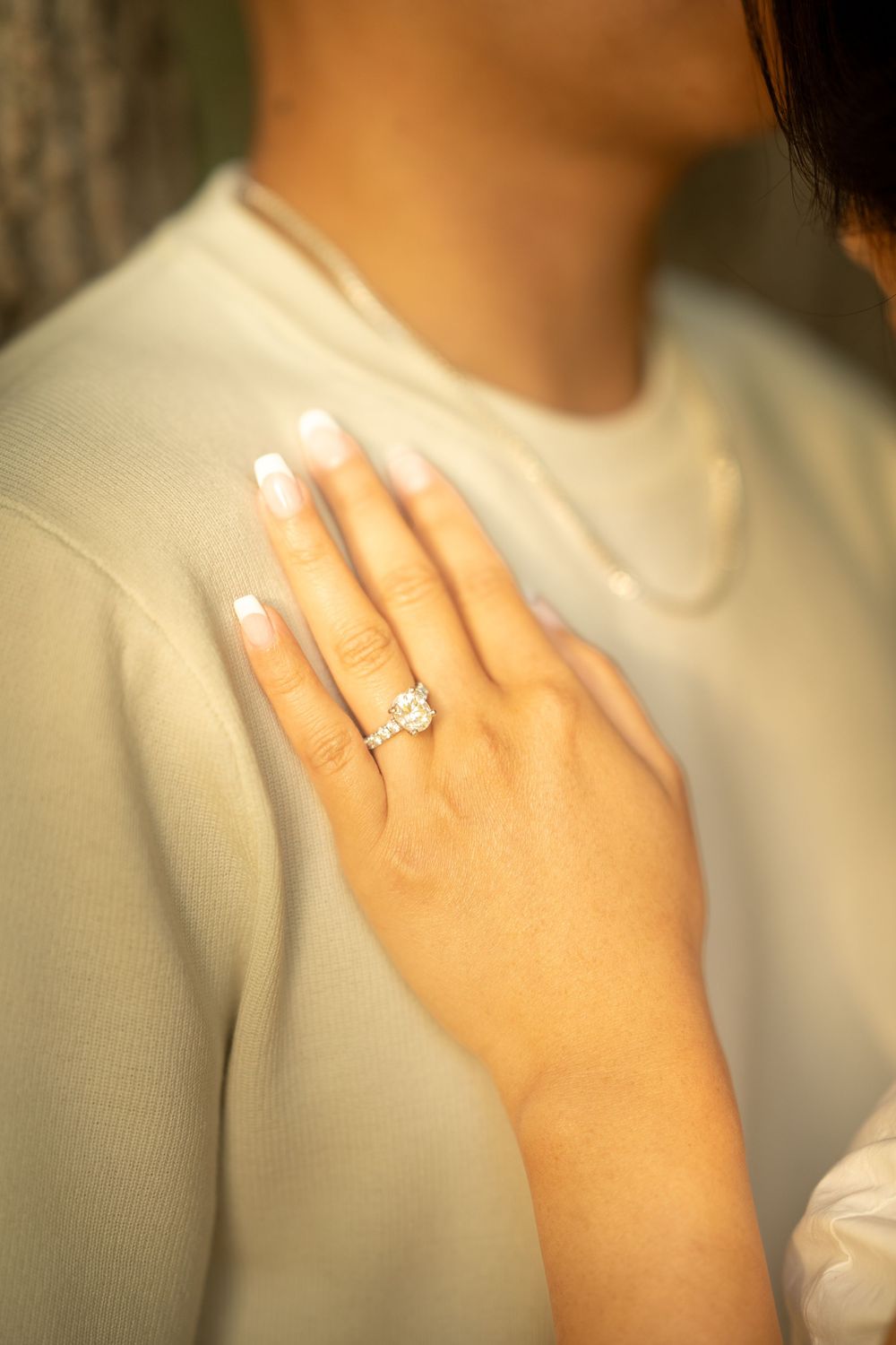 Close-up of a woman's hand displaying a sparkling diamond engagement ring, resting on a man's chest in a white sweater.