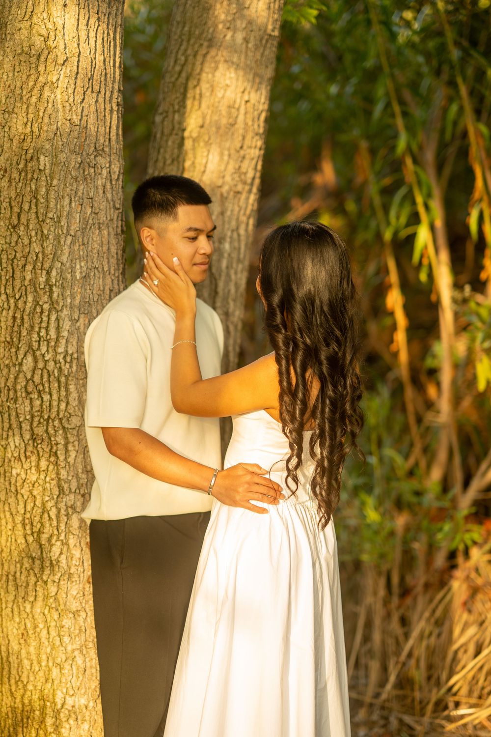 Couple sharing a tender moment near trees, woman in white dress touching mans face in golden sunset light.