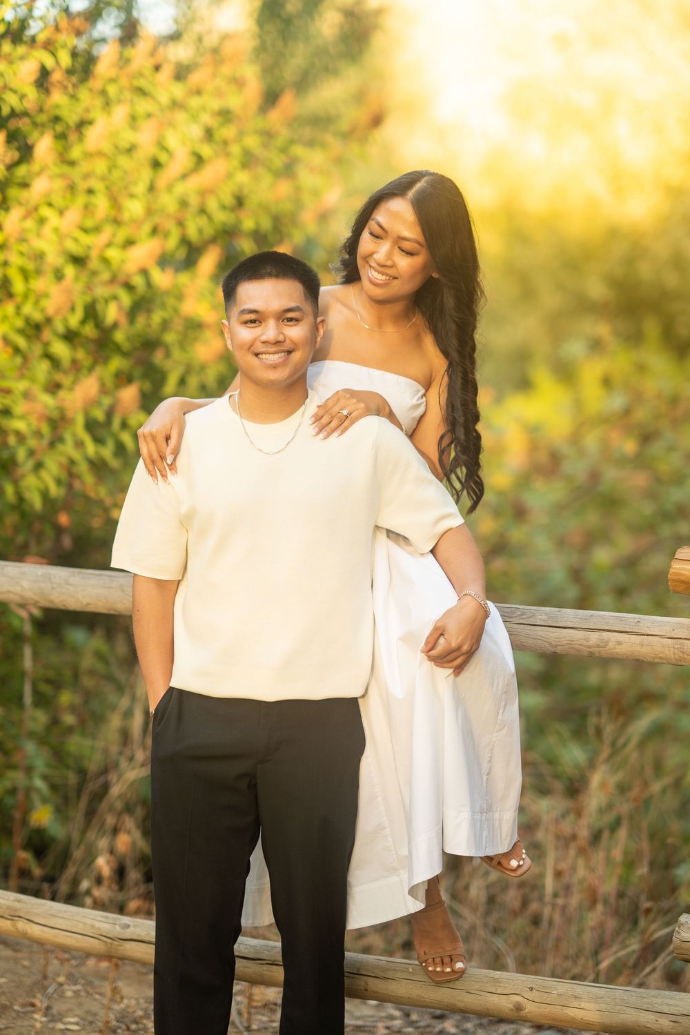 Young couple posing outdoors at golden hour, woman in white dress embracing man in cream shirt by wooden fence.