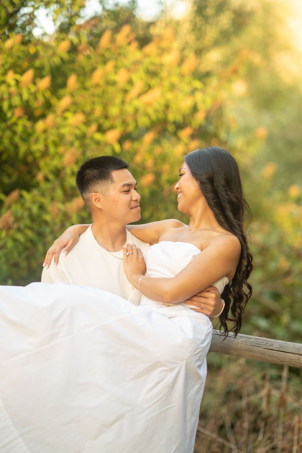 Man carrying woman in white wedding dress outdoors, surrounded by autumn foliage in warm golden light.