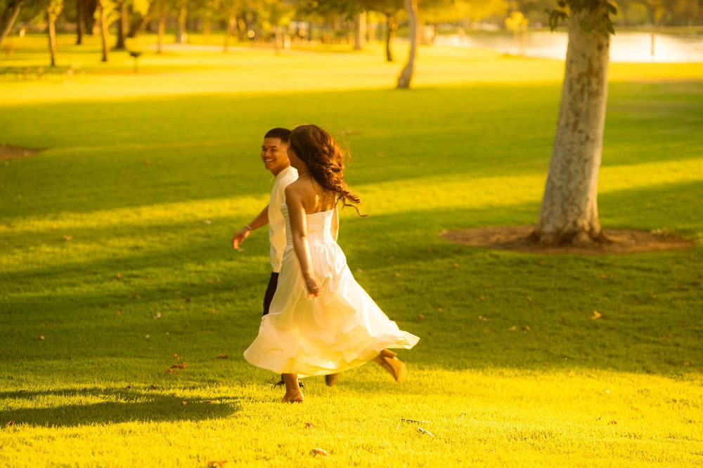 A couple in wedding attire runs joyfully across lush green grass in a sunlit park.