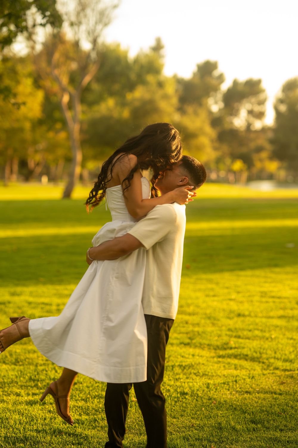 Man lifting woman in white dress during romantic embrace on green grass at sunset in a park.