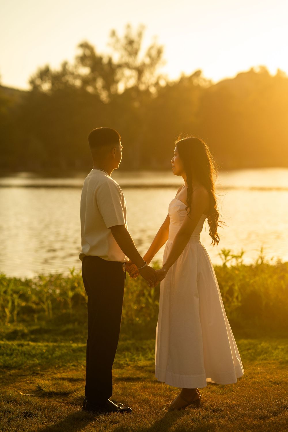 Asian couple holding hands at golden sunset by a lake, woman in white dress and man in casual attire.