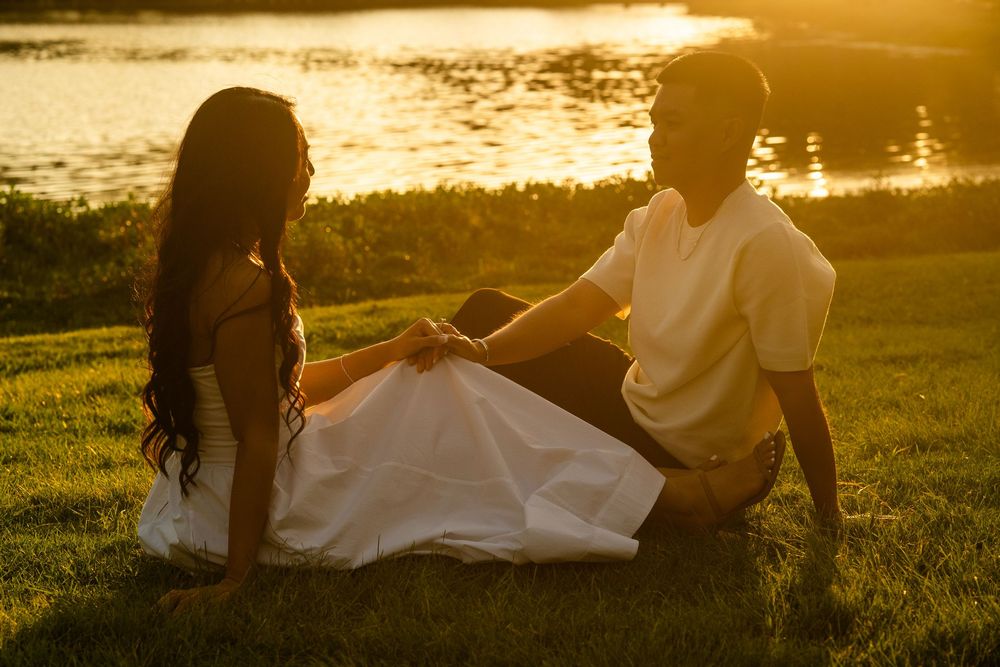 Couple sitting together on grass by a lake at sunset, holding hands in warm golden evening light.