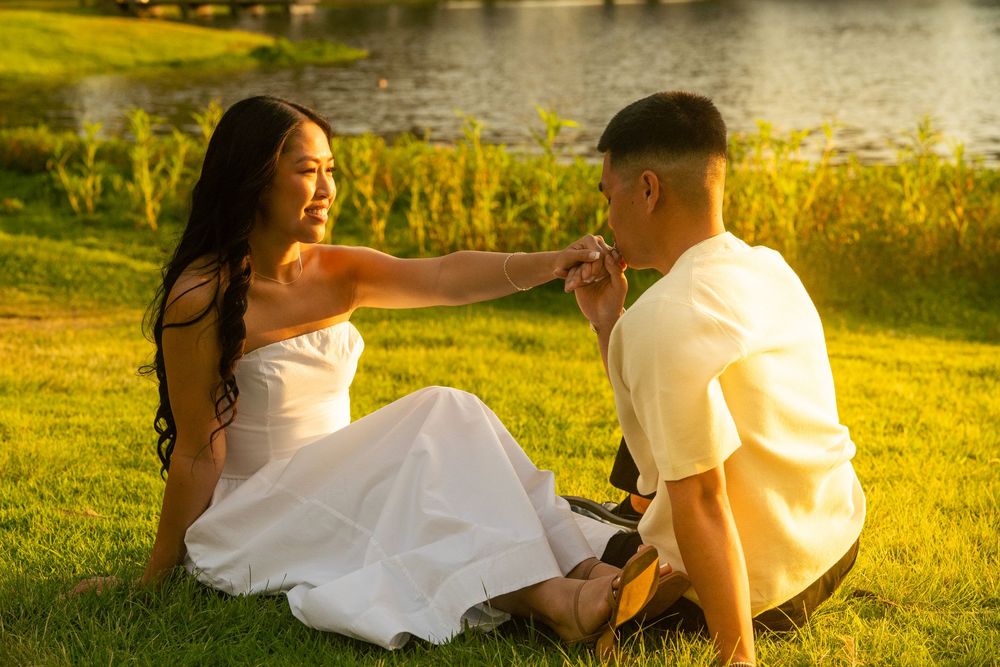 Couple sitting on grass by lake at sunset, woman in white dress holding man's hand romantically.