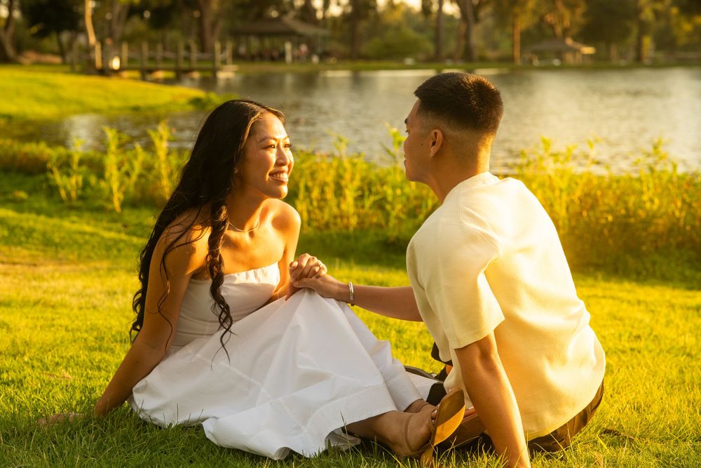 Couple sitting on grass by a sunlit lake, holding hands and gazing at each other during golden hour.