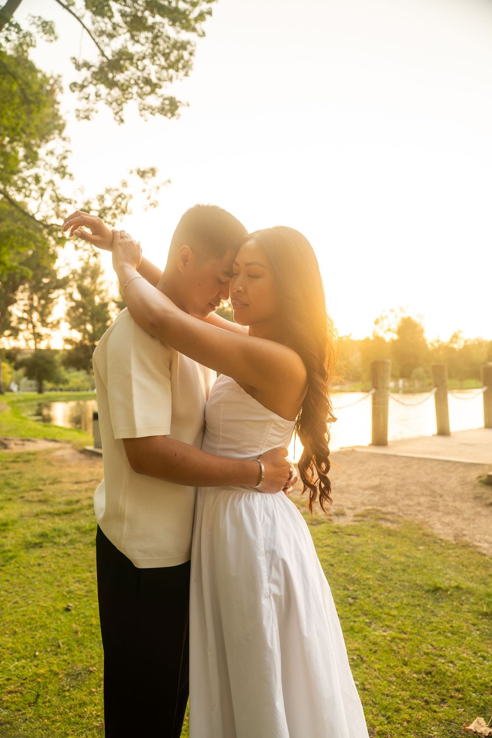 Couple dancing and embracing near a lake at sunset, bathed in warm golden light among lush trees.