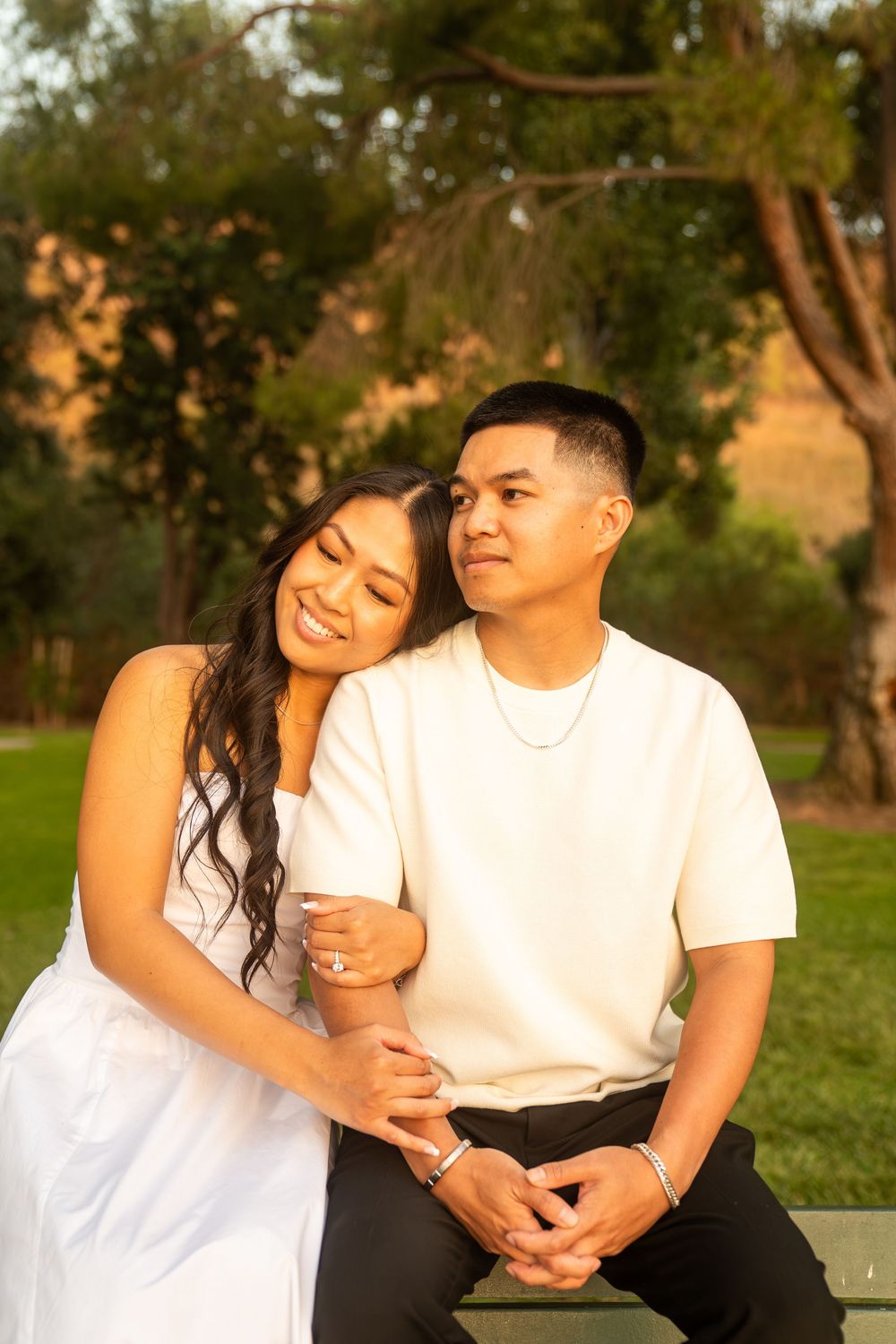 Happy young couple sitting together in a park at golden hour, woman in white dress leaning on mans shoulder smiling.