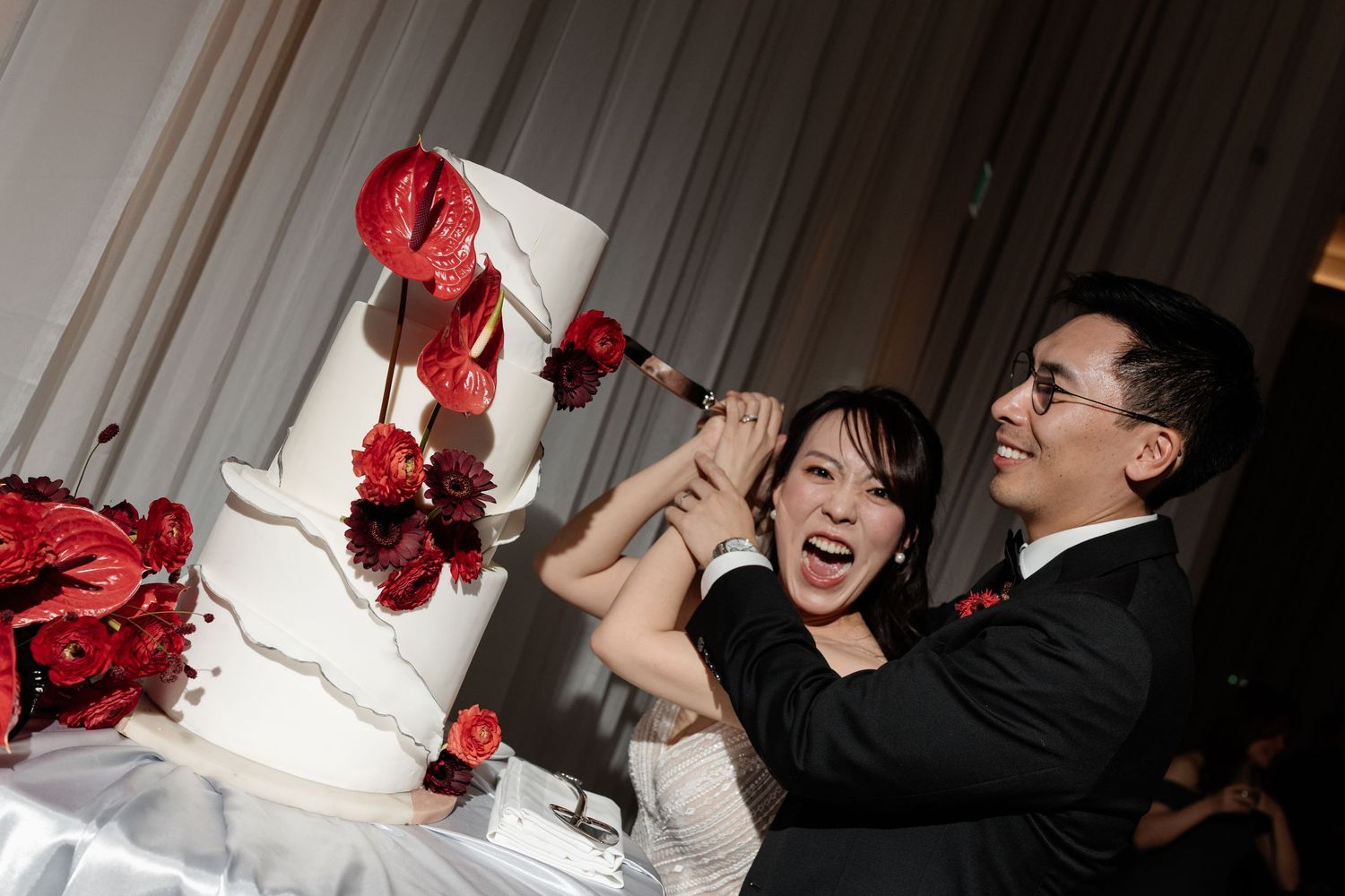 Joyful bride and groom cutting a white wedding cake decorated with red anthurium flowers and roses.