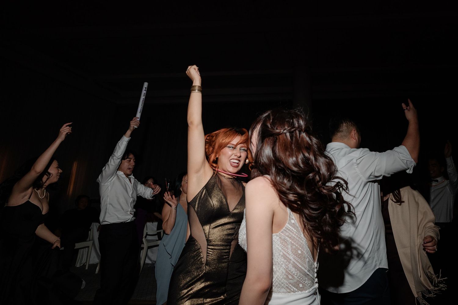 Guests dancing joyfully at a wedding reception, with a redhead in a gold dress raising her fist in celebration.