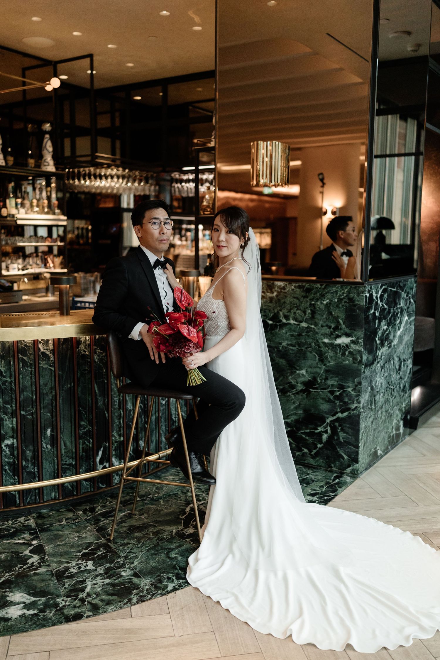 Elegant bride in white gown holding red bouquet poses with groom in tuxedo at a marble bar venue.