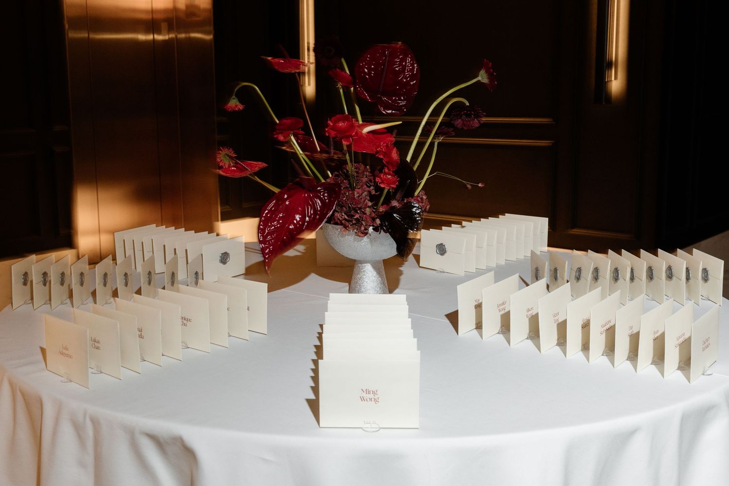 Wedding escort card table with red anthurium floral centerpiece surrounded by neatly arranged place cards.