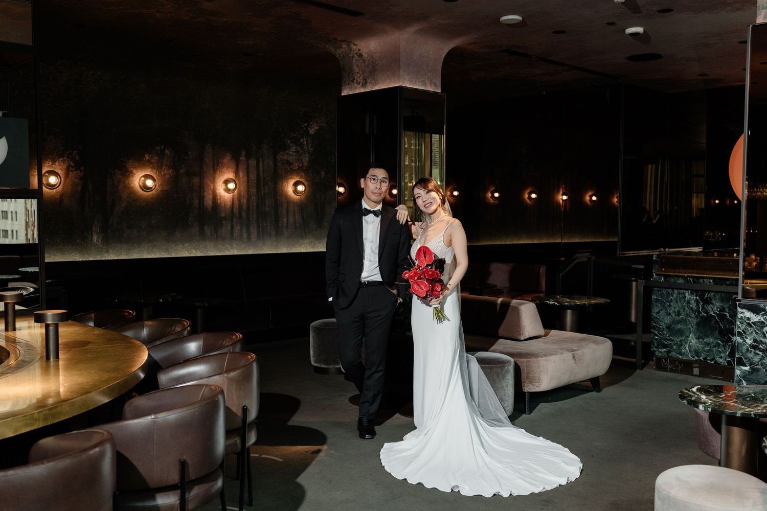 Bride and groom pose together in a moody dark luxury bar venue holding a red bridal bouquet.