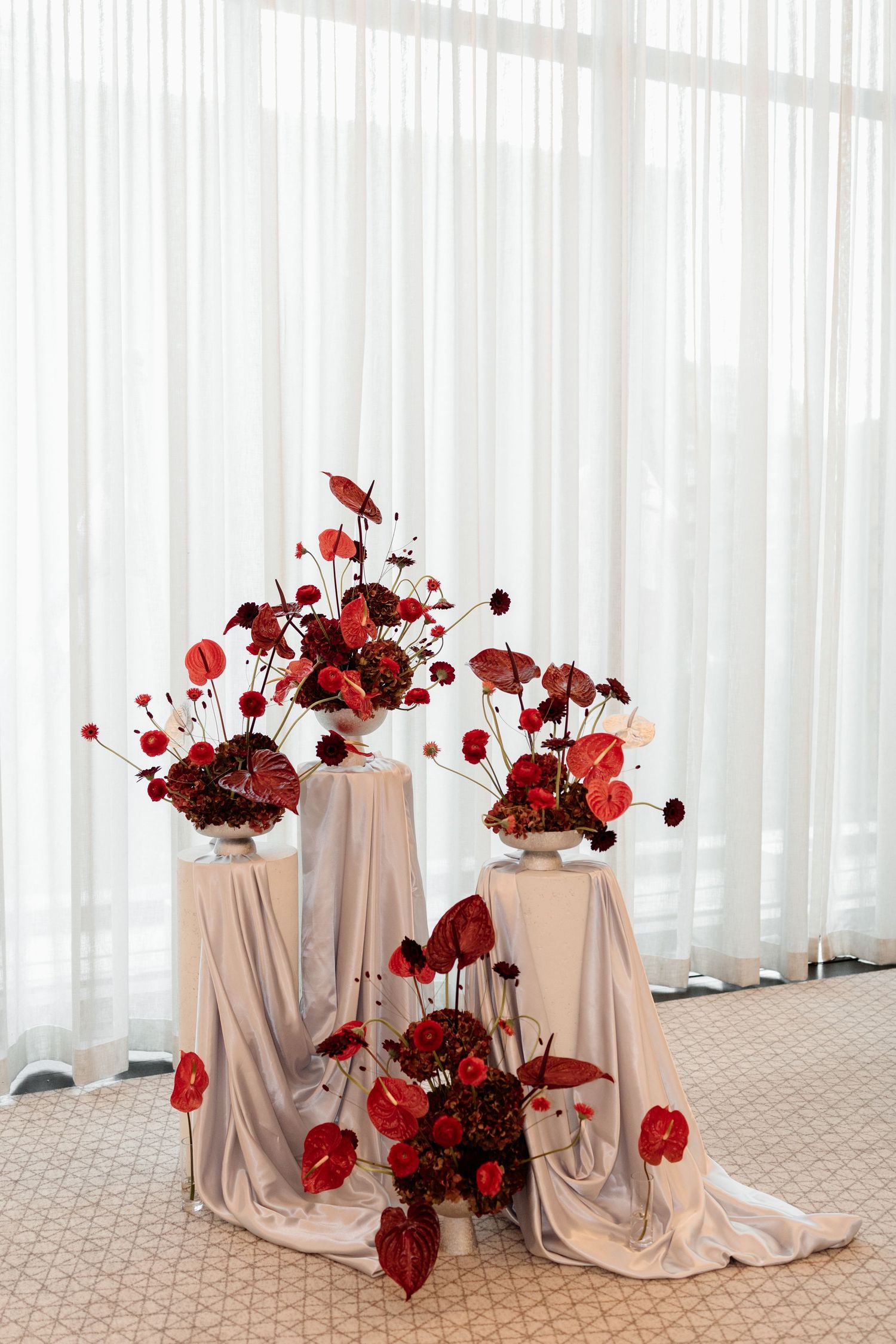 Three elegant floral arrangements with red anthuriums and roses on draped white pedestals against sheer curtains.