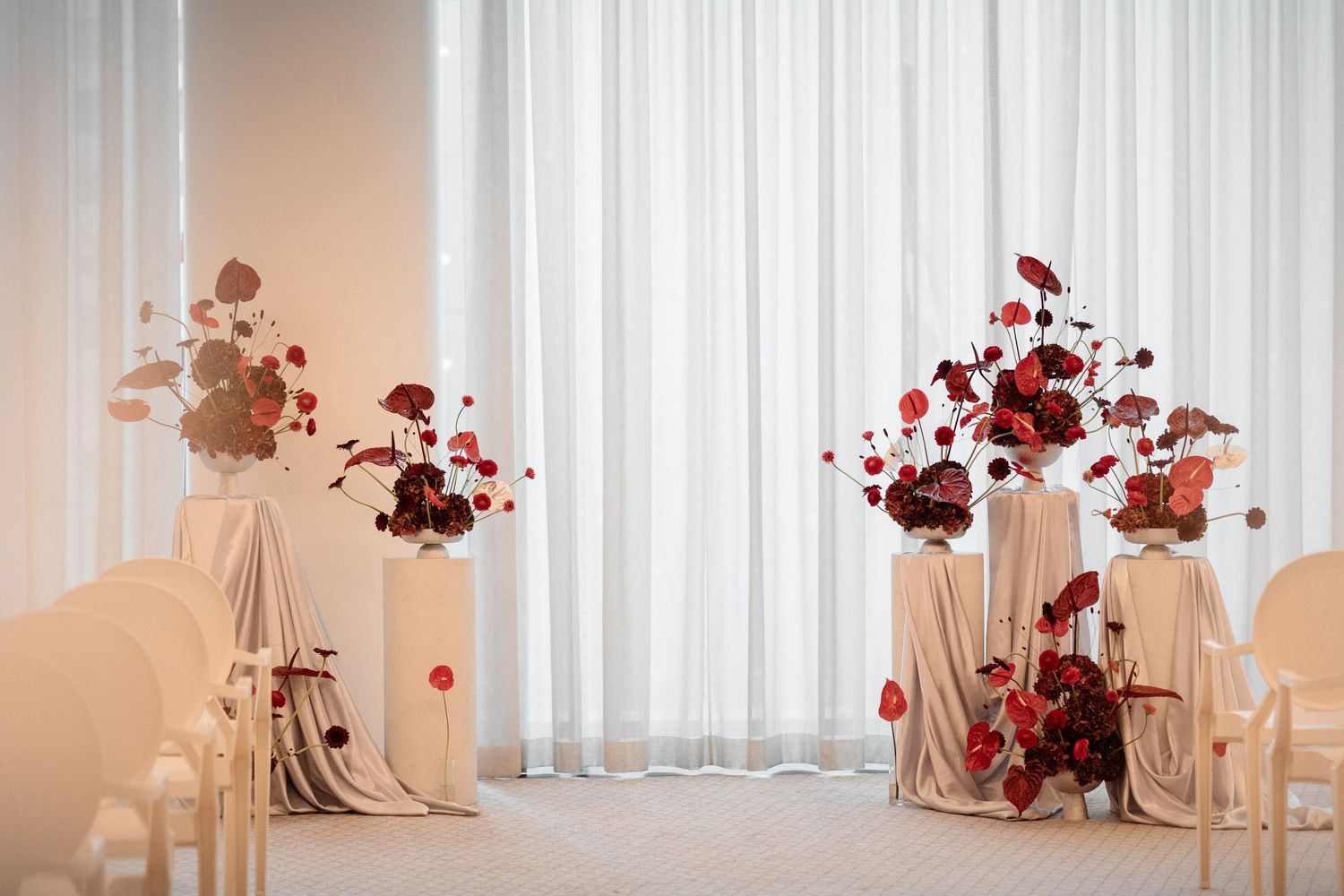 Elegant wedding ceremony setup with red floral arrangements on draped tables against sheer white curtains.