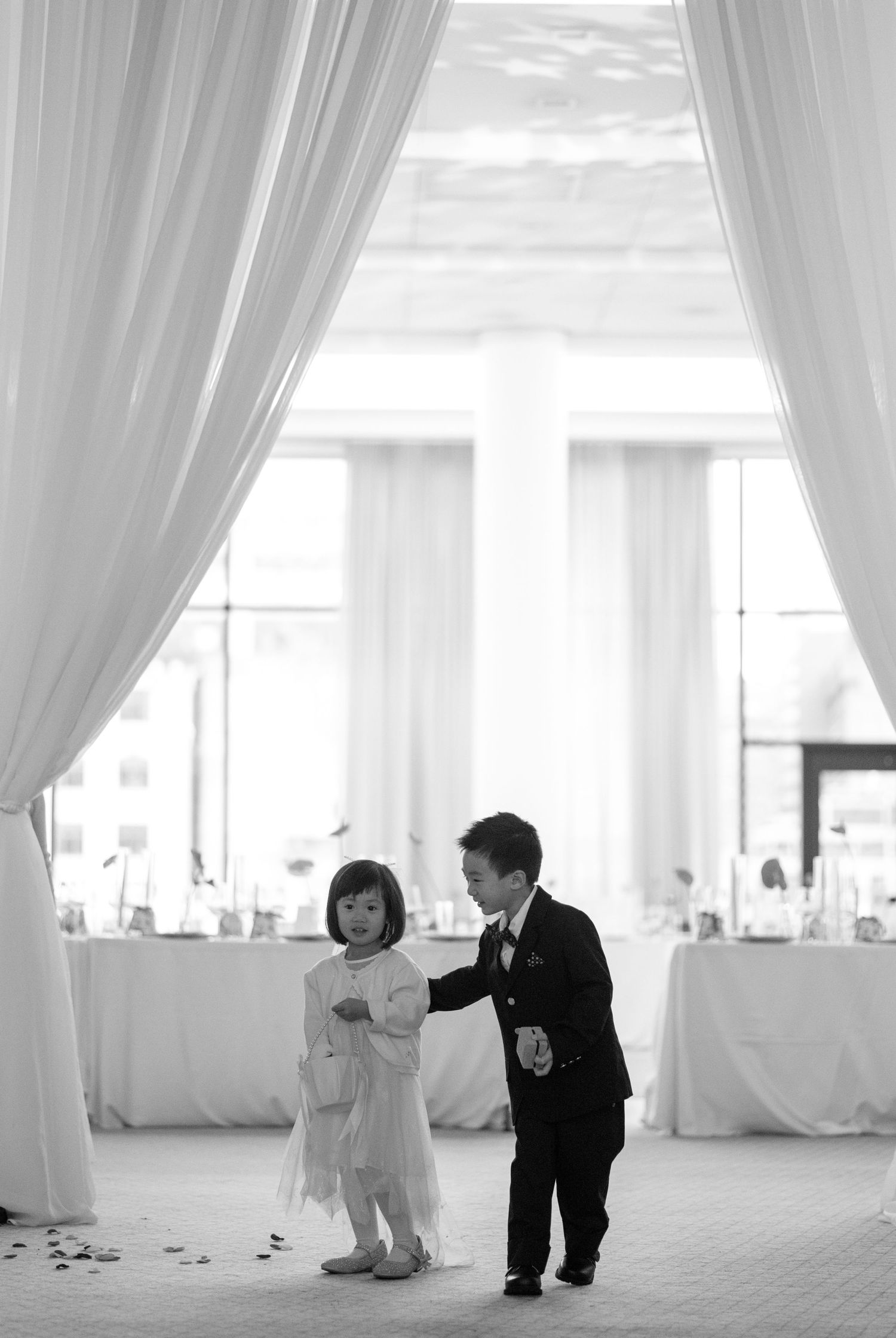 Black and white photo of two young children, a flower girl and ring bearer, standing together in a wedding venue.
