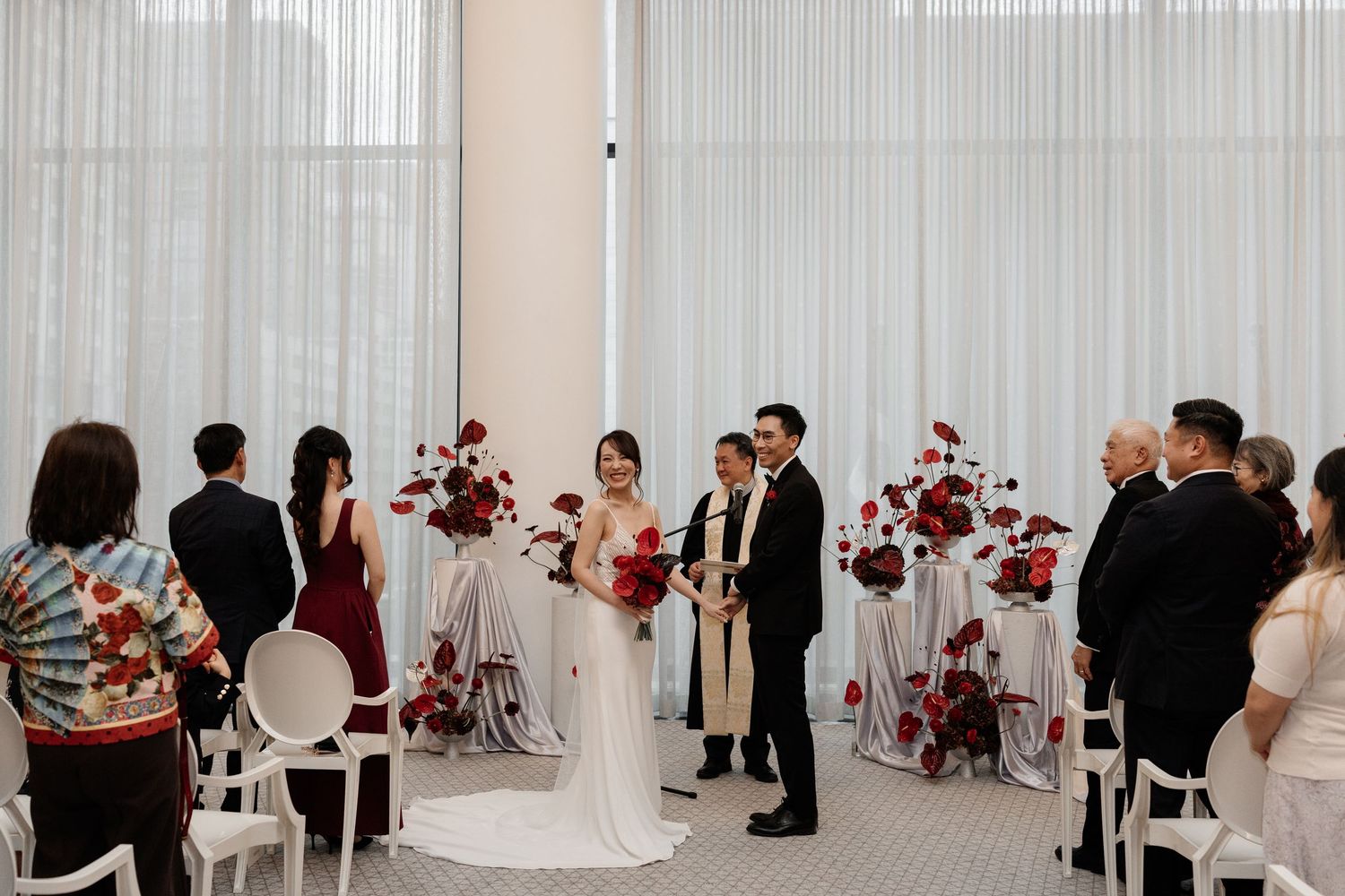 Elegant wedding ceremony with couple at altar, red floral arrangements, white chairs, and sheer curtain backdrop.