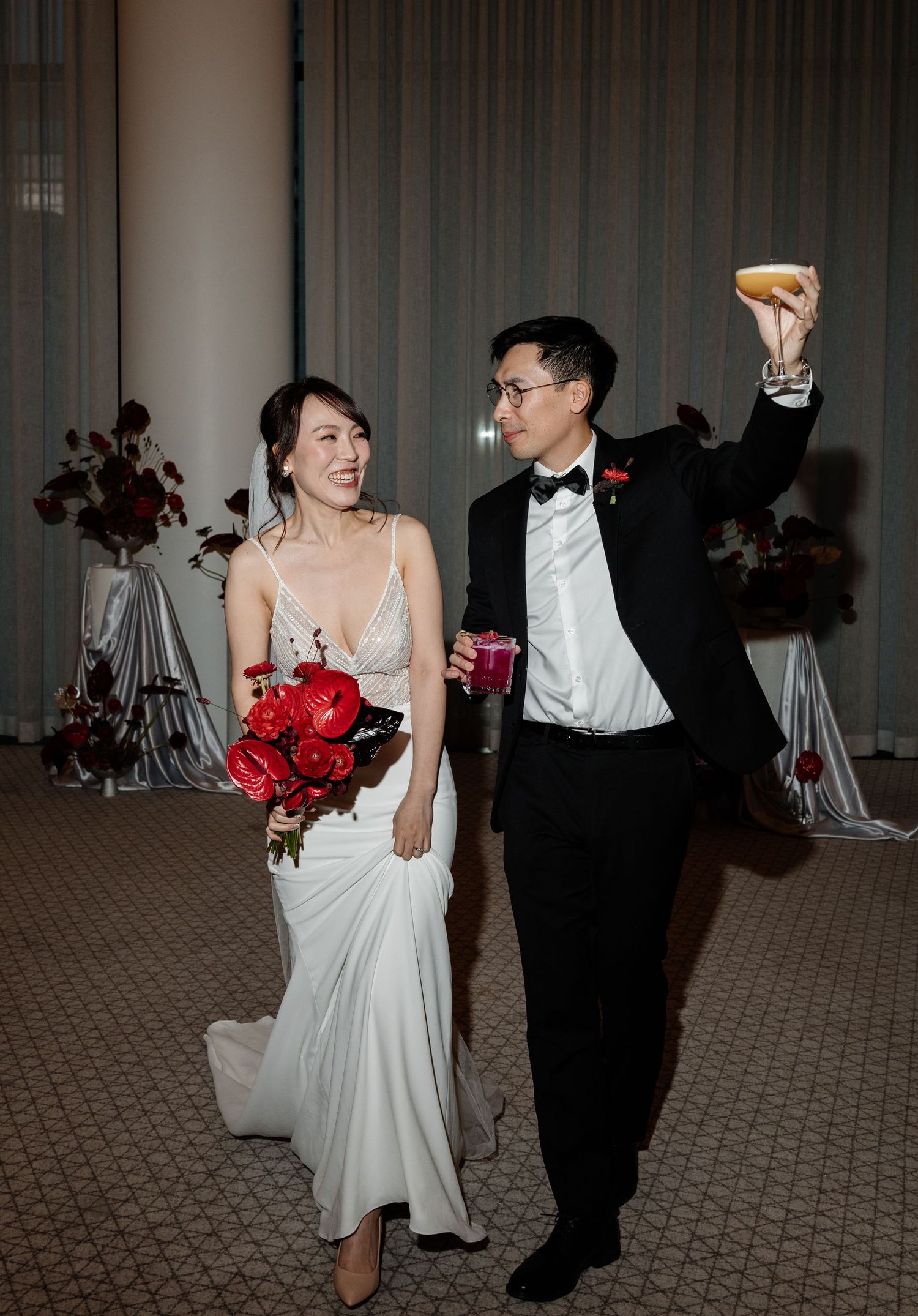 Bride in white gown holding red bouquet stands with groom in tuxedo raising a drink at their wedding reception.