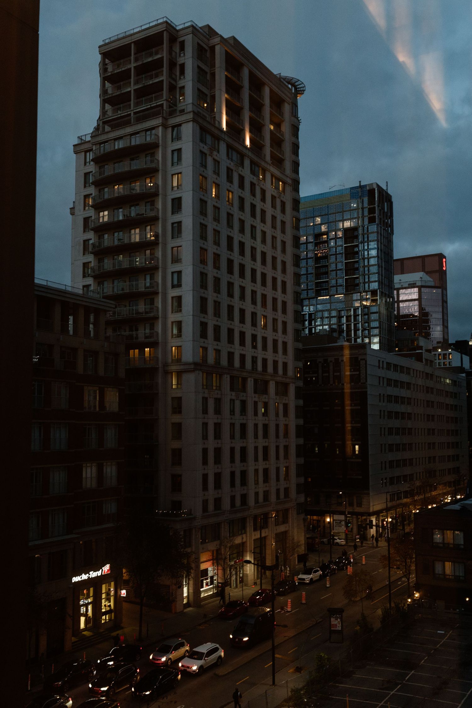 Twilight cityscape view of illuminated downtown buildings and street traffic under a cloudy evening sky.