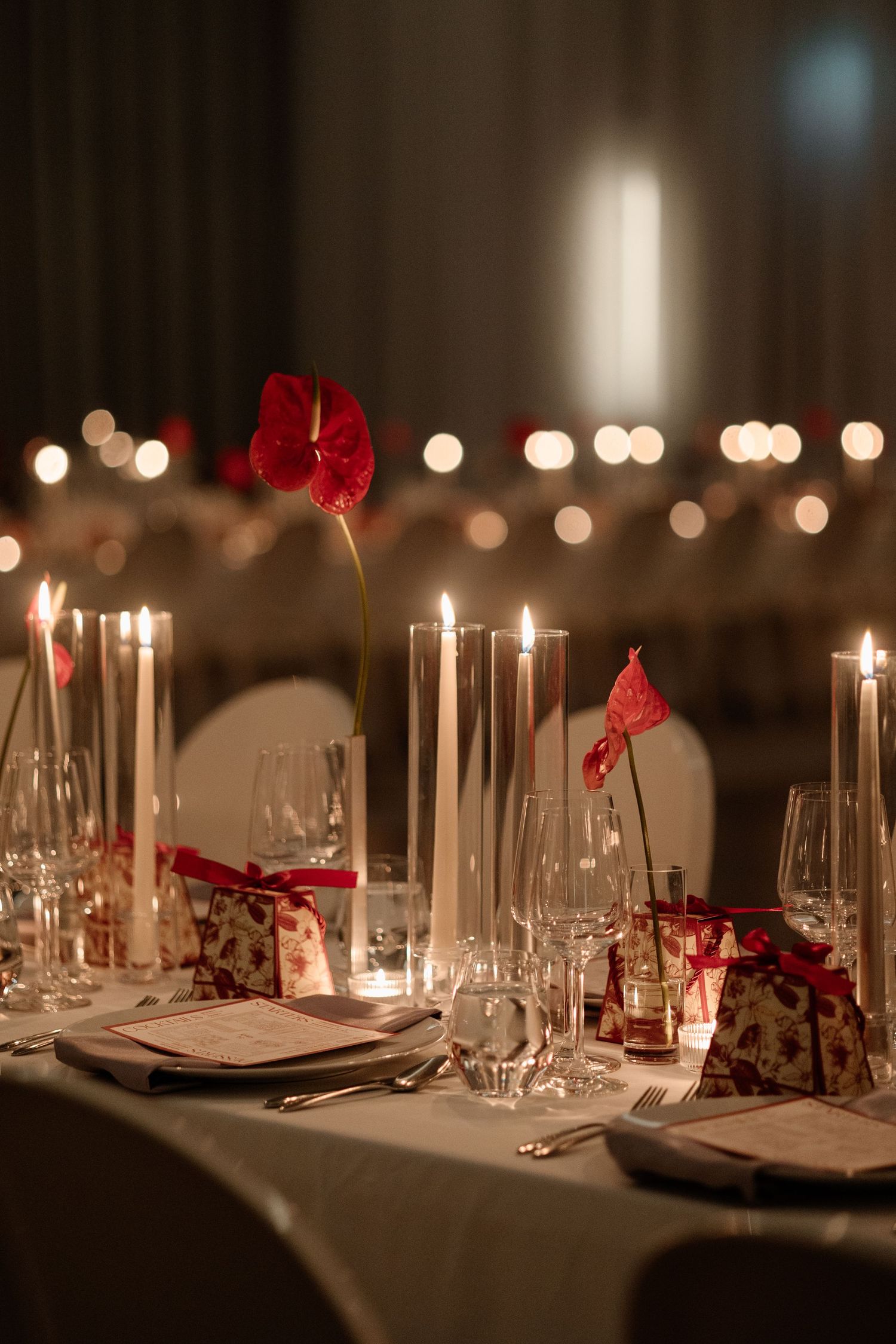 Elegant wedding reception table with red anthurium flowers, tall white candles, crystal glassware, and warm bokeh lighting.