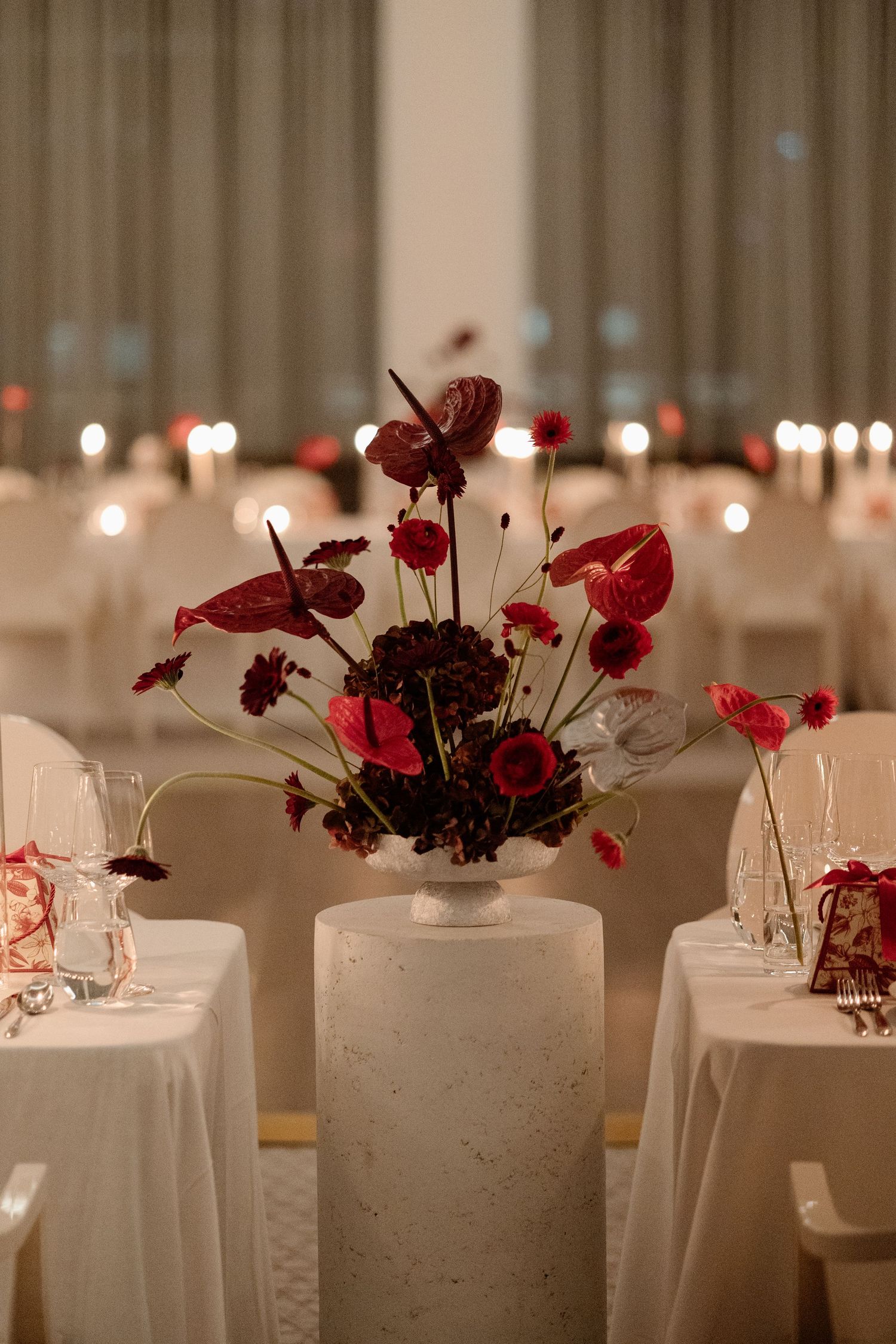Elegant wedding reception centerpiece with deep red anthuriums and burgundy flowers in a white stone vase.