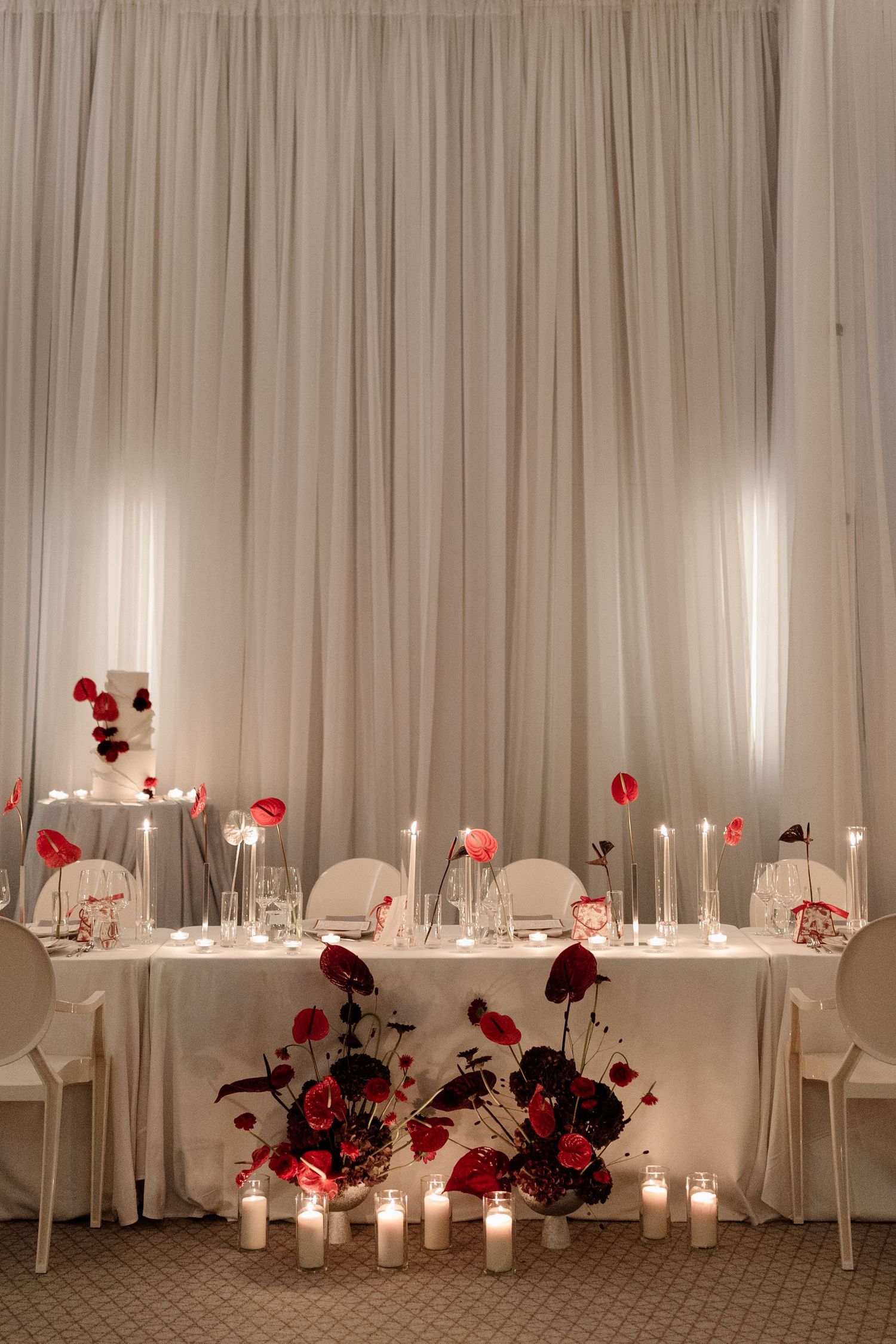 Romantic wedding sweetheart table with red roses, tall candles, and white drape backdrop decor.