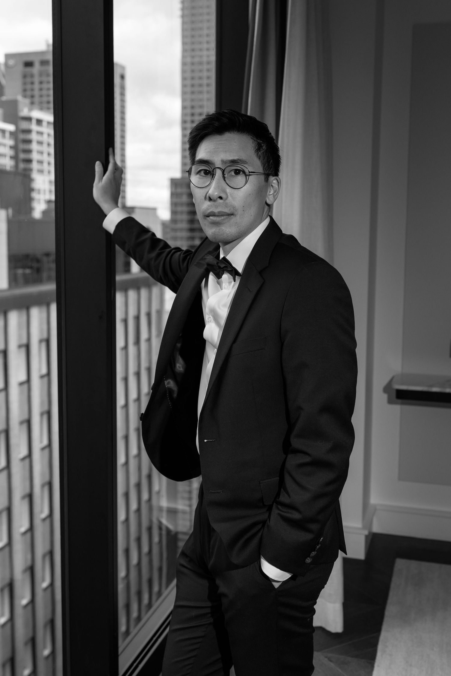 Man in black tuxedo and bow tie with glasses posing by a window with city skyline in black and white.