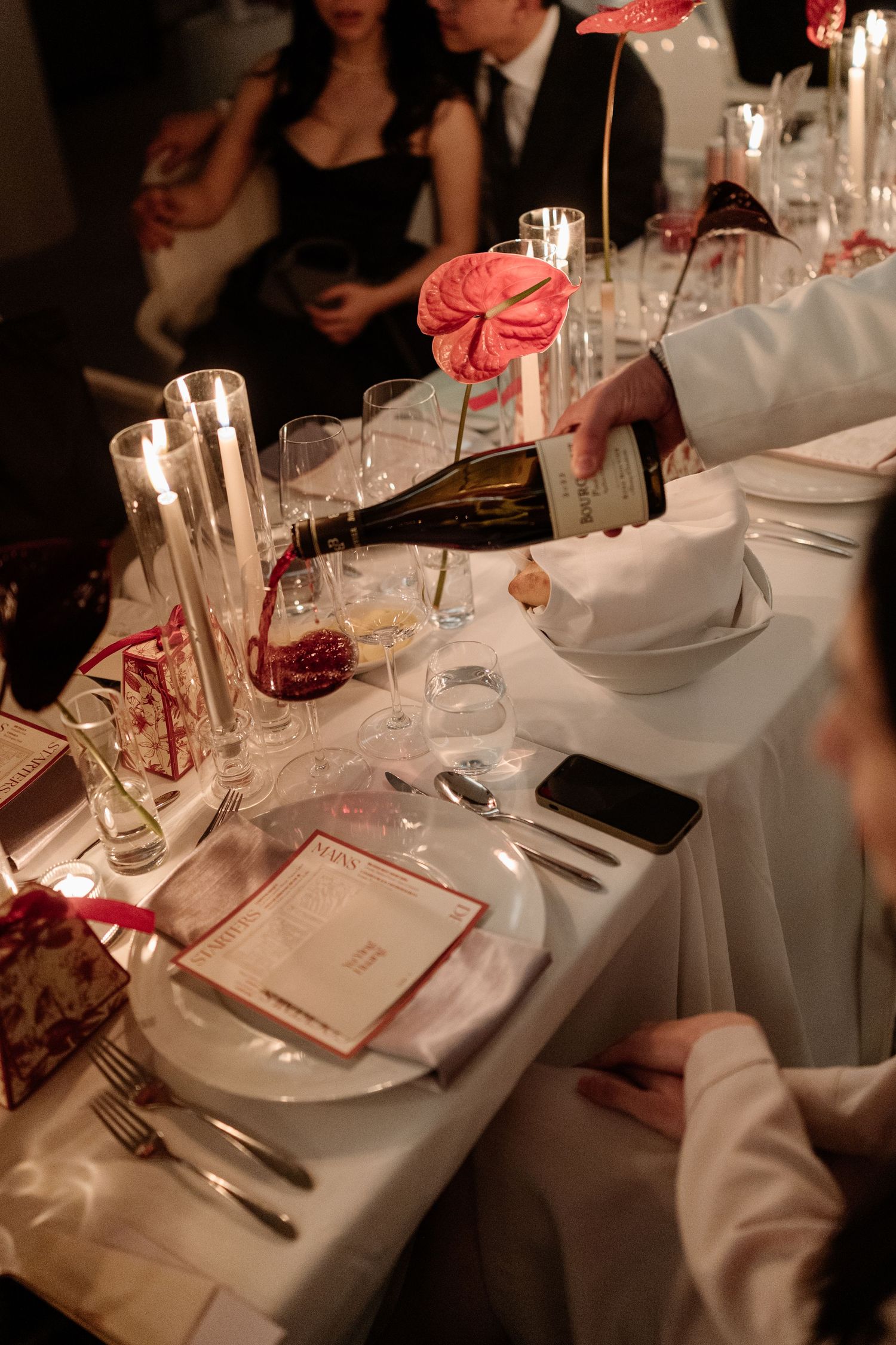 Waiter pouring red wine at an elegantly set dining table with candles, glassware, and pink floral decorations.