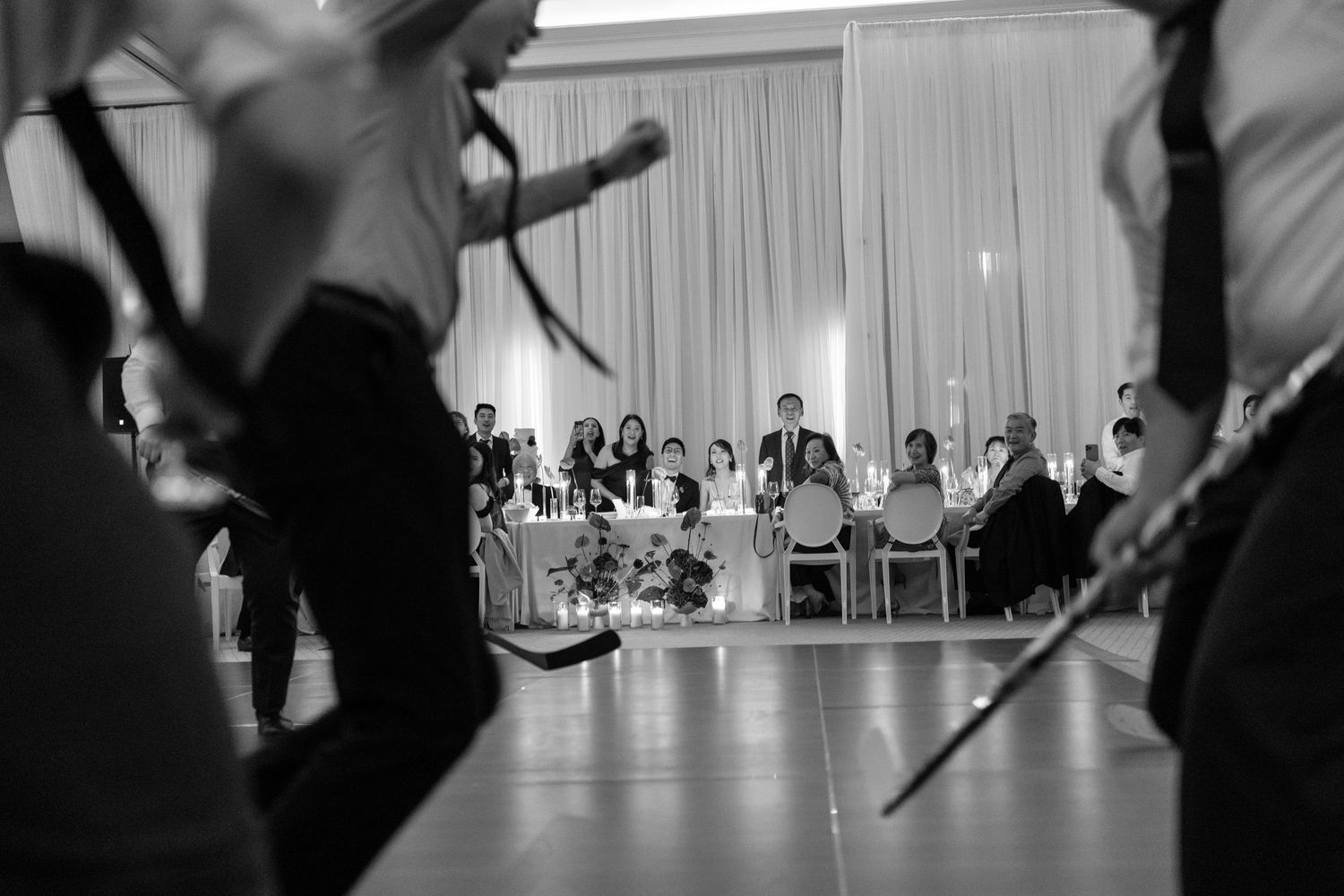 Black and white wedding reception photo of traditional sword dancers performing before seated guests at elegant venue.