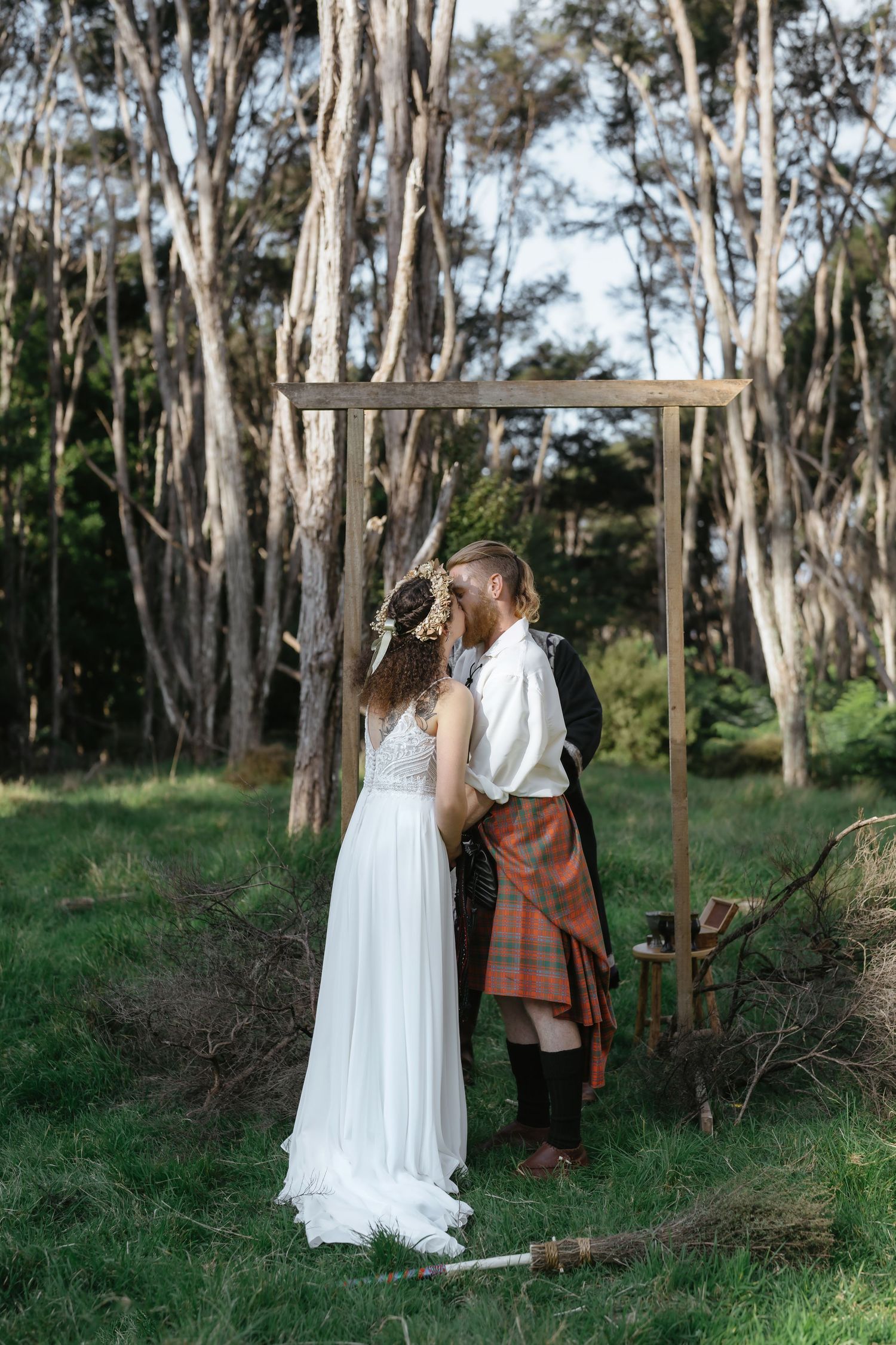 Bride and groom sharing a kiss under a wooden arch in a lush forest, groom wearing a traditional Scottish kilt.
